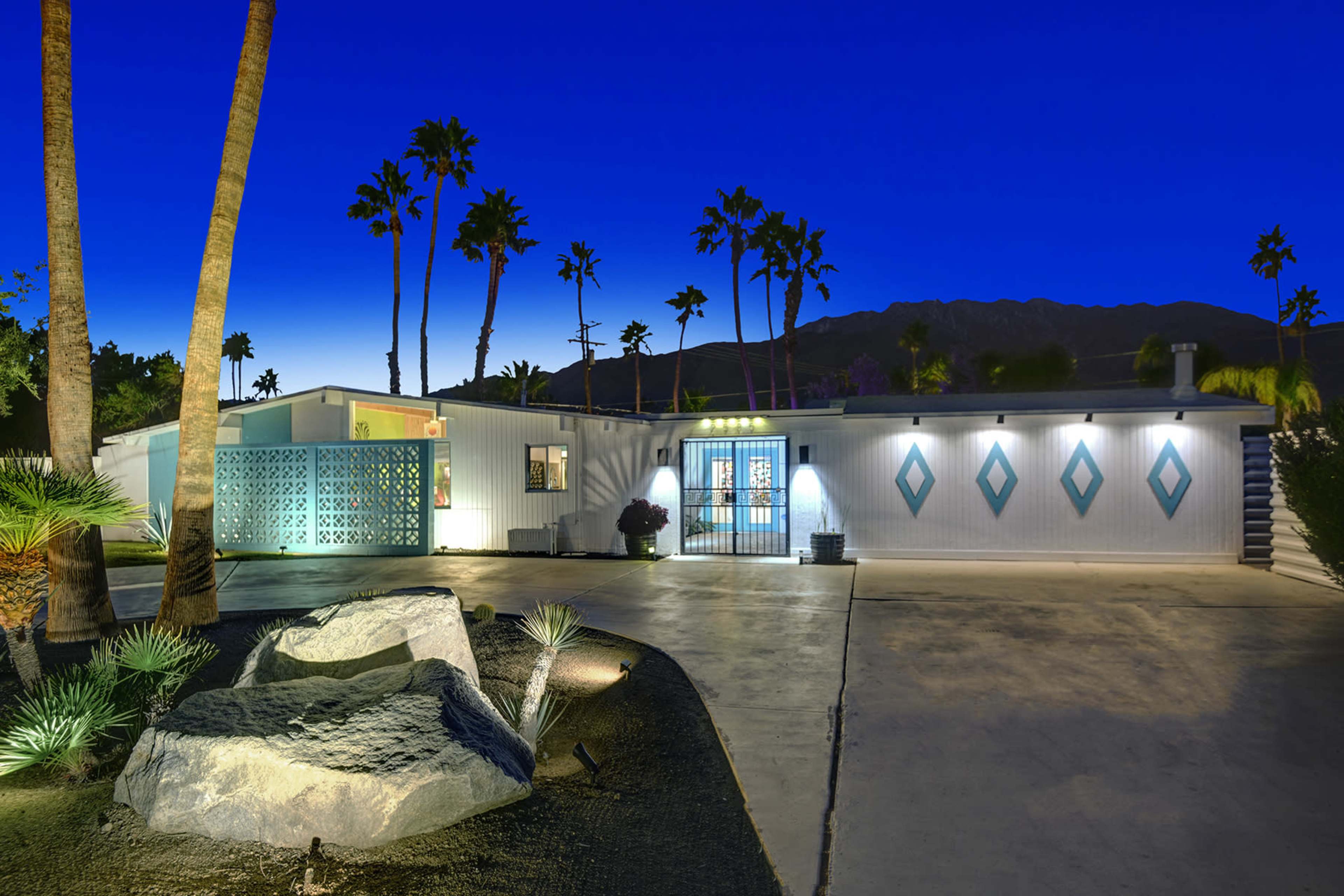 The image shows a modern, single-story home with a landscaped yard, palm trees, and a mountain backdrop under a twilight sky.