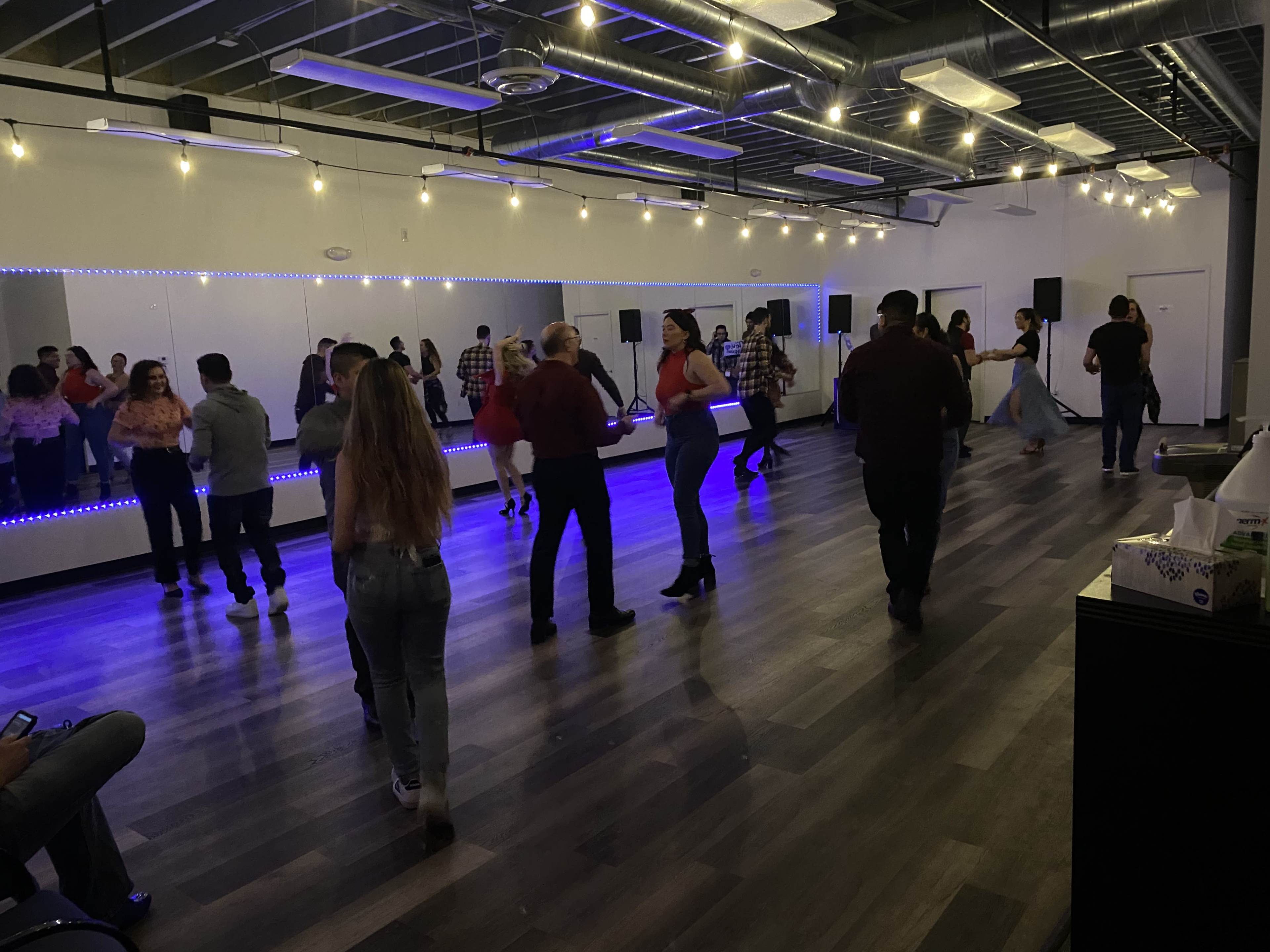 A group of people are dancing in a spacious room with mirrored walls and colorful lighting.