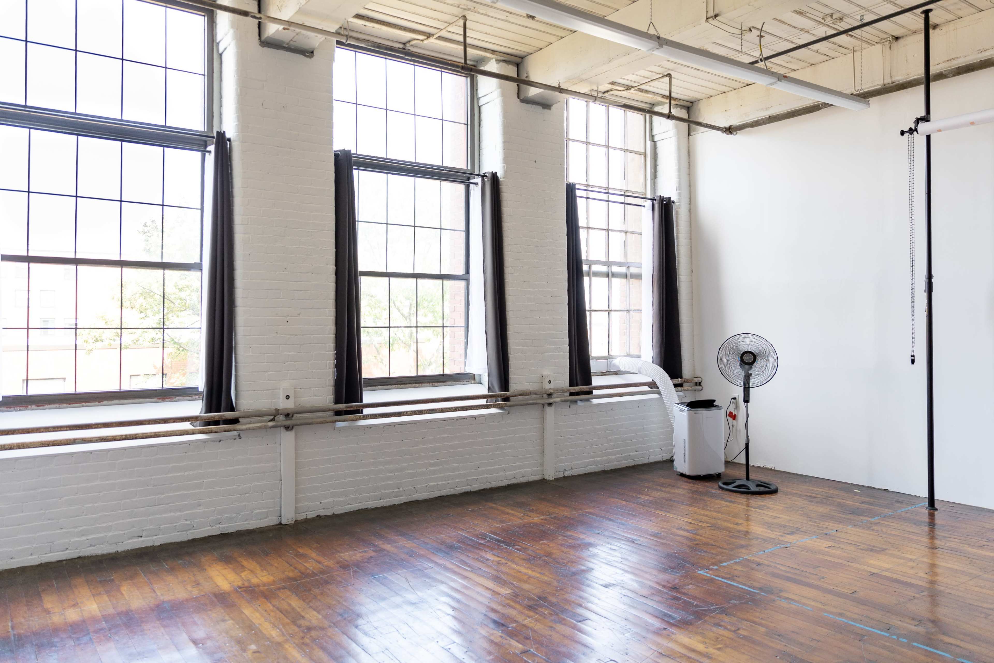 A spacious room with large windows, wooden flooring, a standing fan, and a white wall.