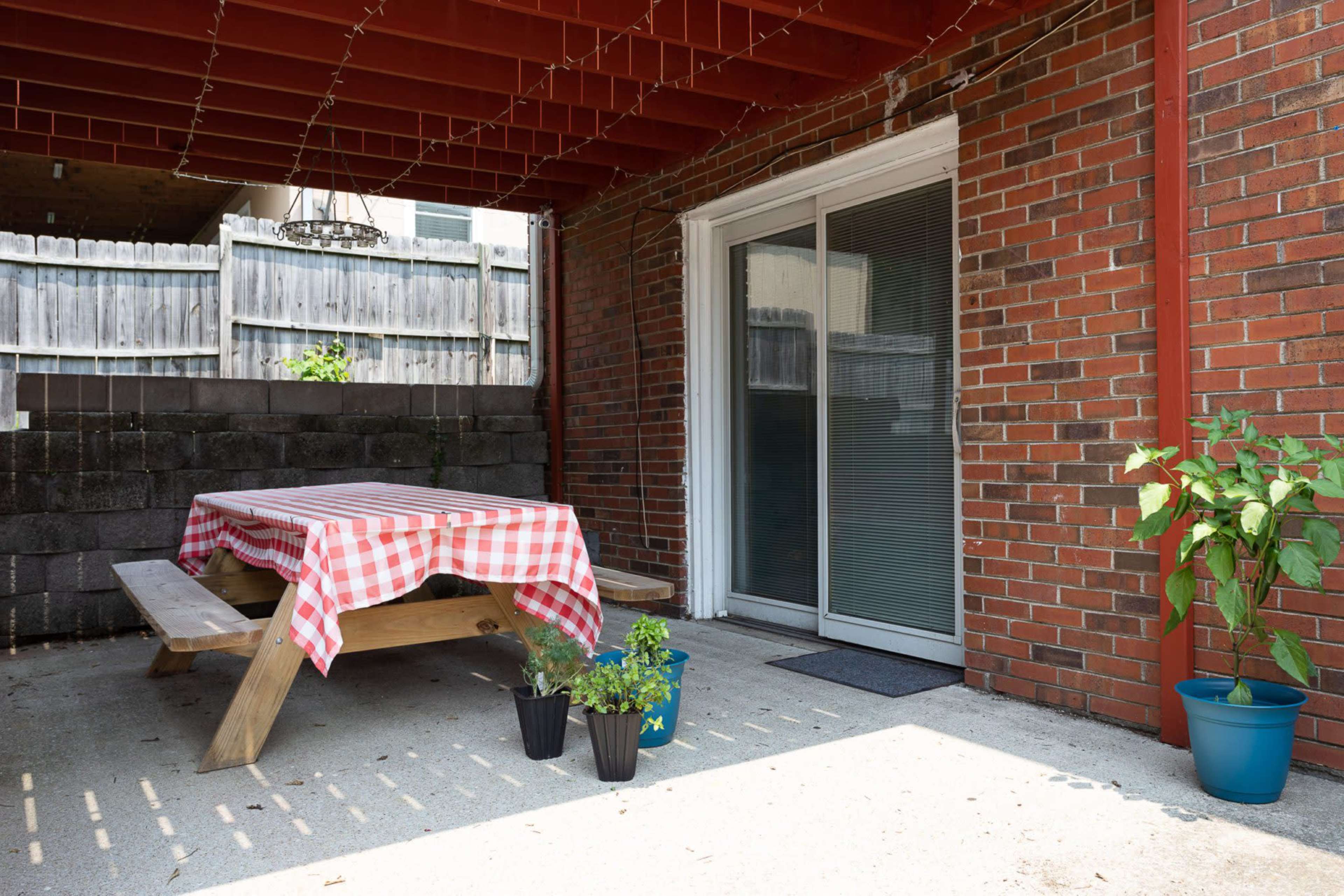 A covered patio features a wooden picnic table with a checkered tablecloth, flanked by two potted plants and sliding glass doors.