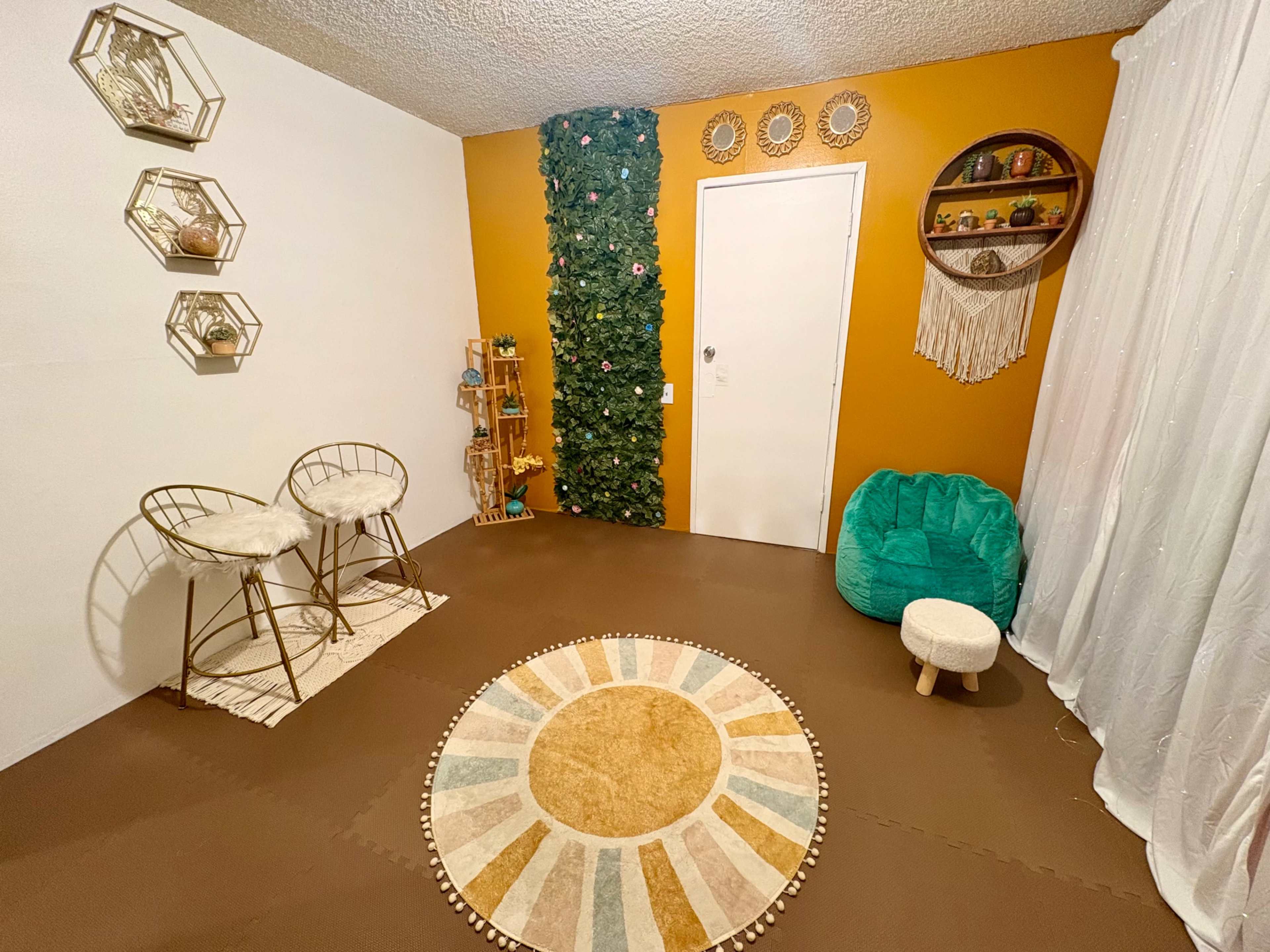 The image shows a cozy, modern room with two gold bar stools, a green bean bag chair, a circular area rug, and a decorated wall featuring greenery and decorative shelves.