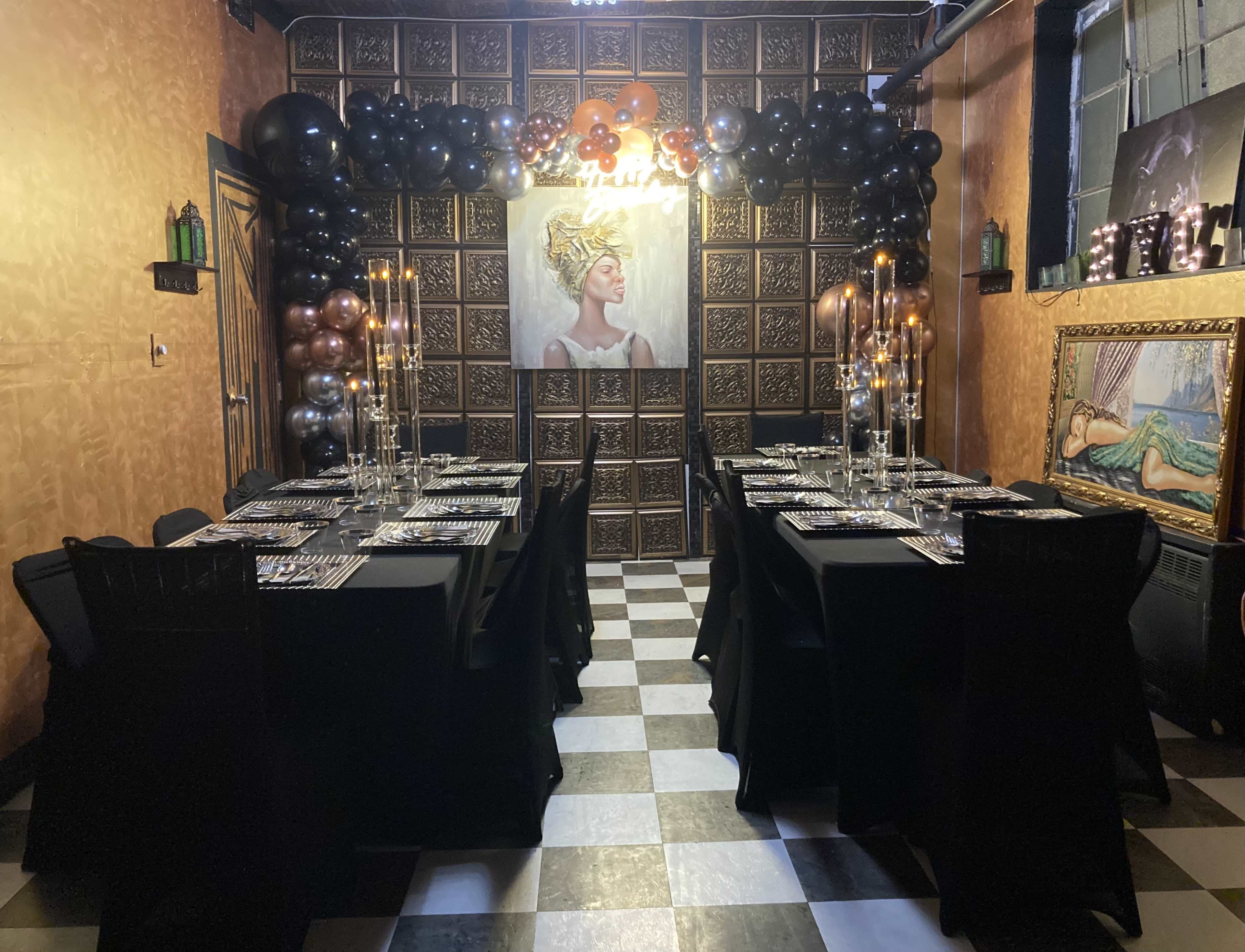 A dining setup features a long table with black tablecloths, elegant place settings, and a backdrop of decorative balloons and a wall art piece in a dimly lit room.