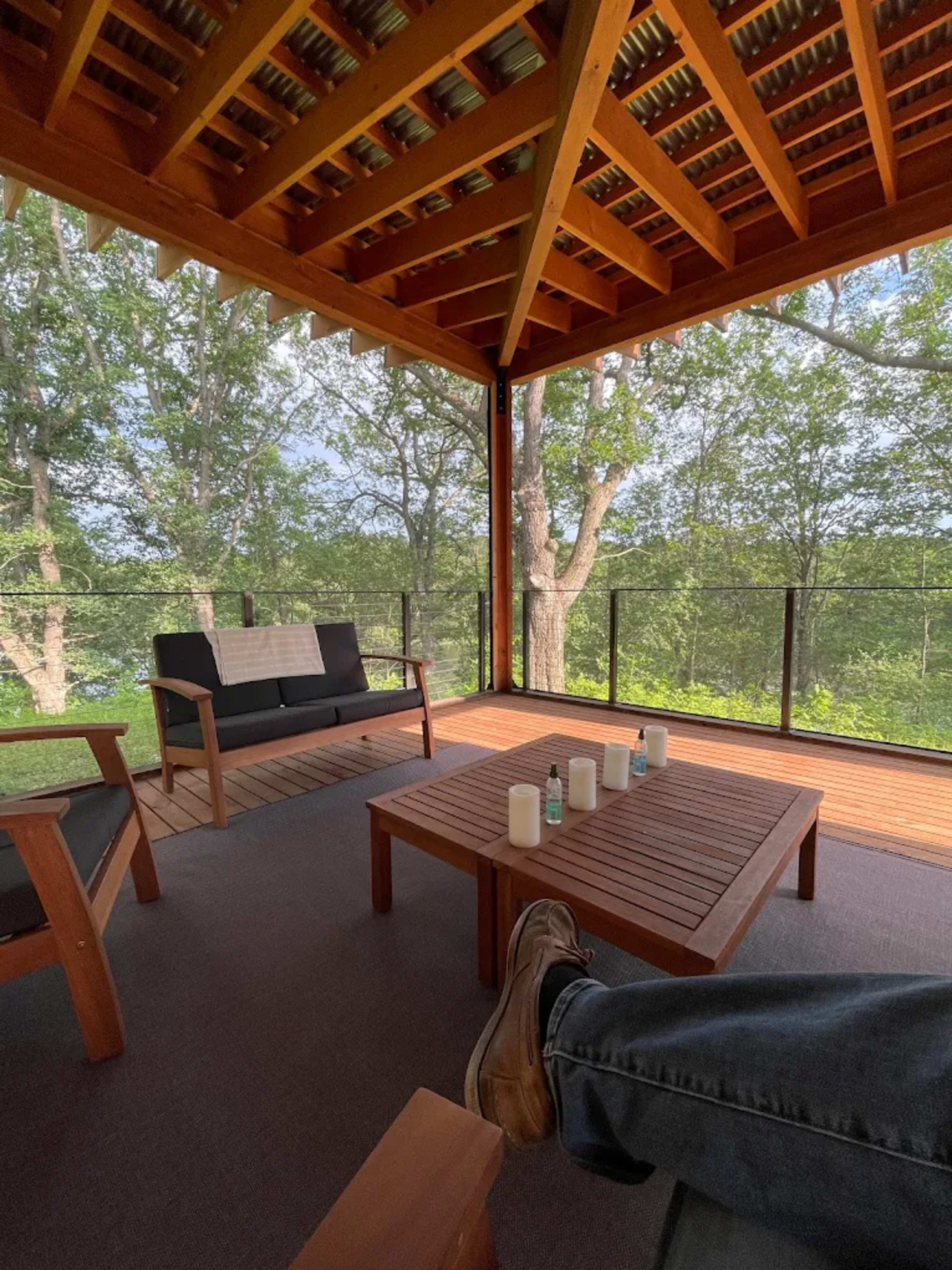 The image shows a wooden patio or balcony with a seating area, a table, and large windows overlooking greenery.