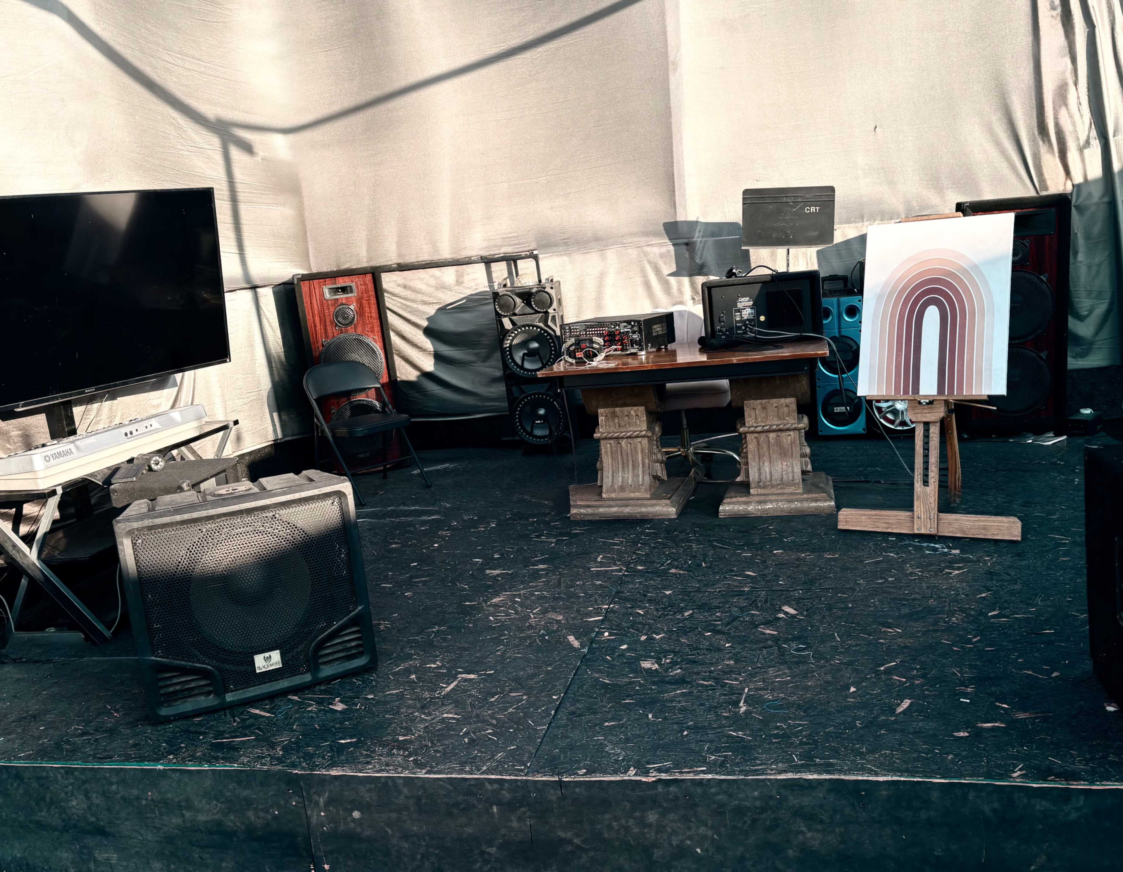 The image shows a sound setup with various speakers, a keyboard, and a piece of artwork displayed on an easel in a tent environment.