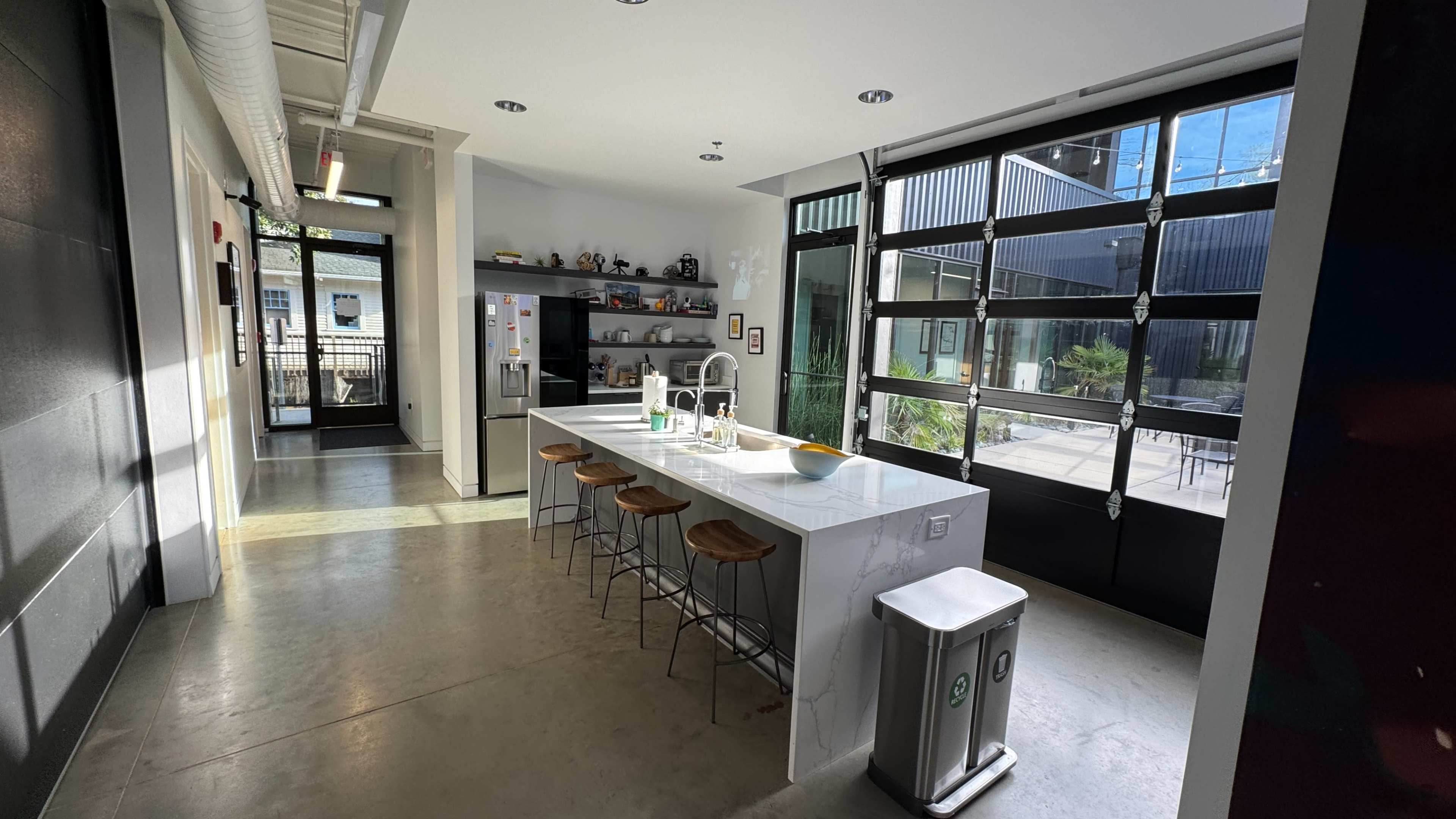 The image shows a modern, open kitchen with a marble island and three wooden stools, alongside large glass doors leading to an outdoor seating area.