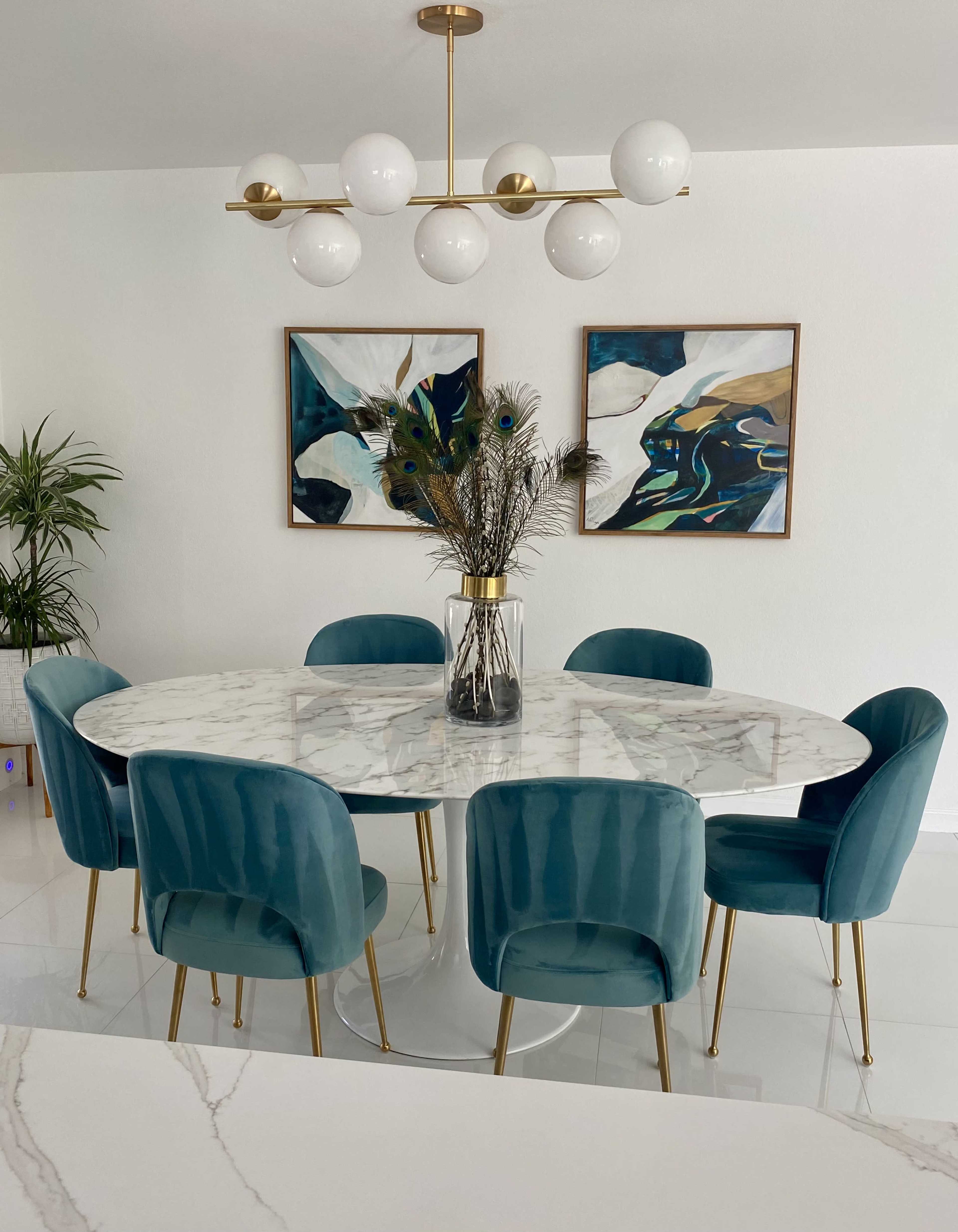 A round marble dining table surrounded by teal upholstered chairs and a floral centerpiece, with abstract artwork hanging on the walls.