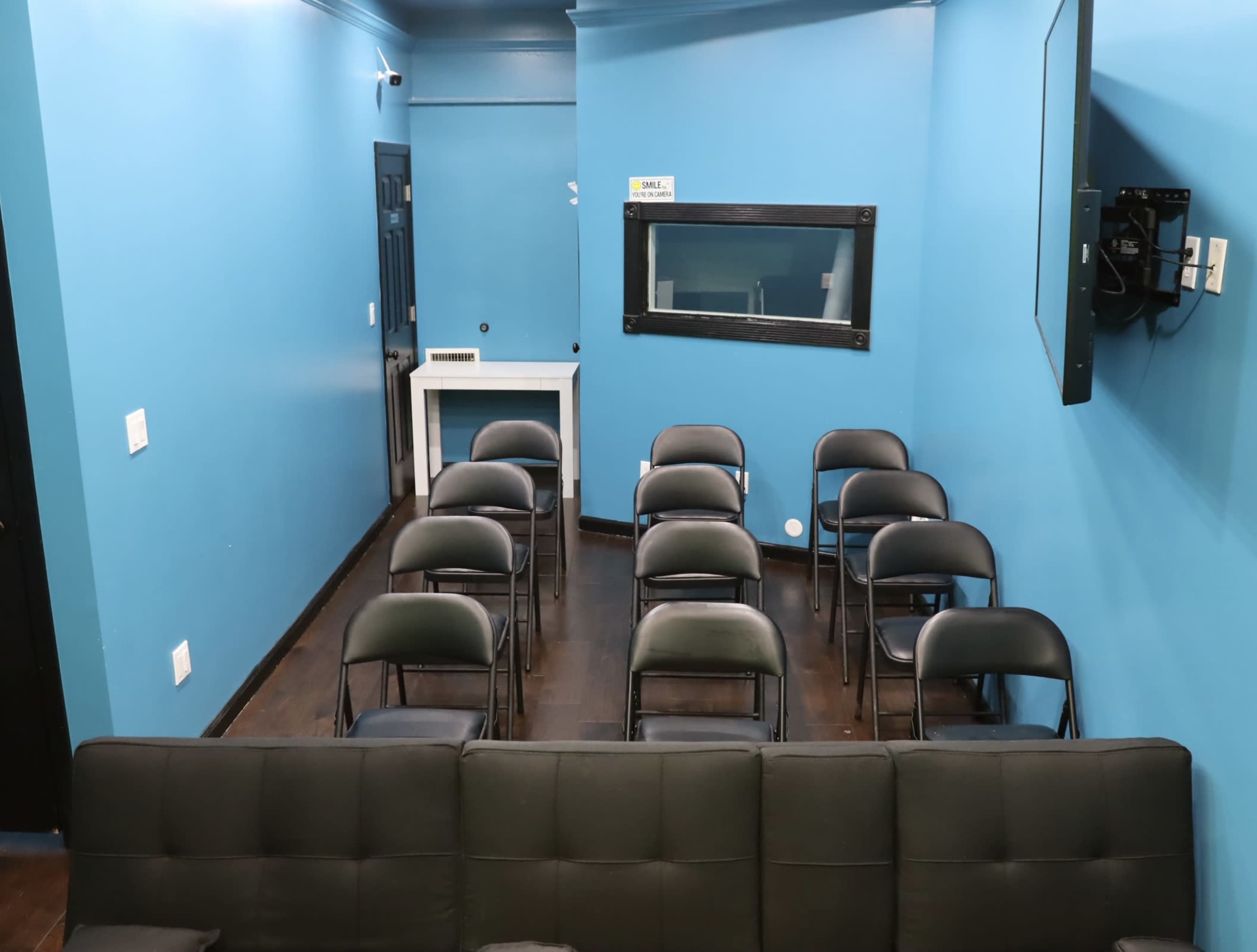 A small room with blue walls, featuring a row of black folding chairs facing a television and a mirrored window.