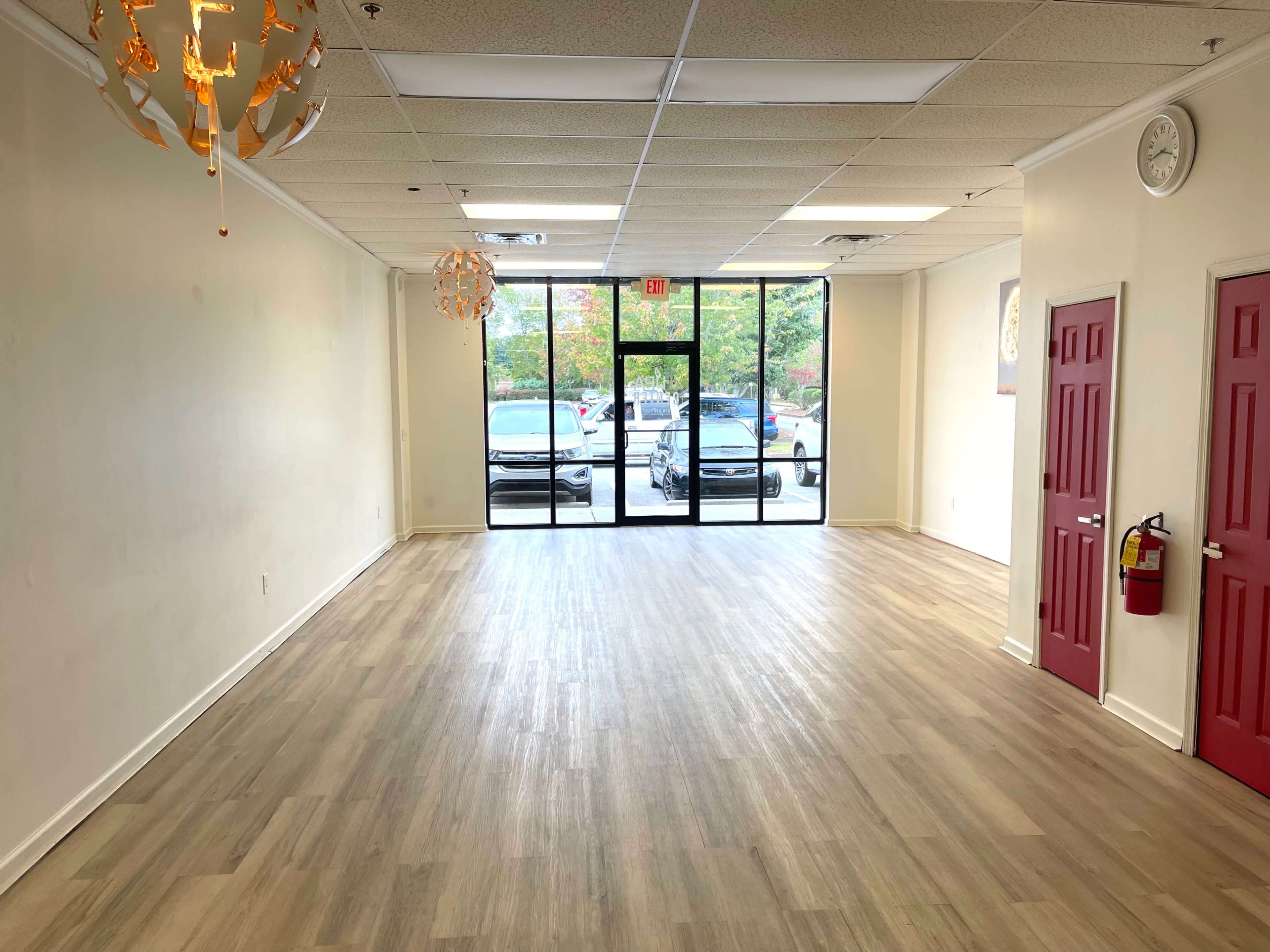 The image shows an empty room with wooden flooring, illuminated by natural light coming through large glass doors that lead outside, which features parked cars.