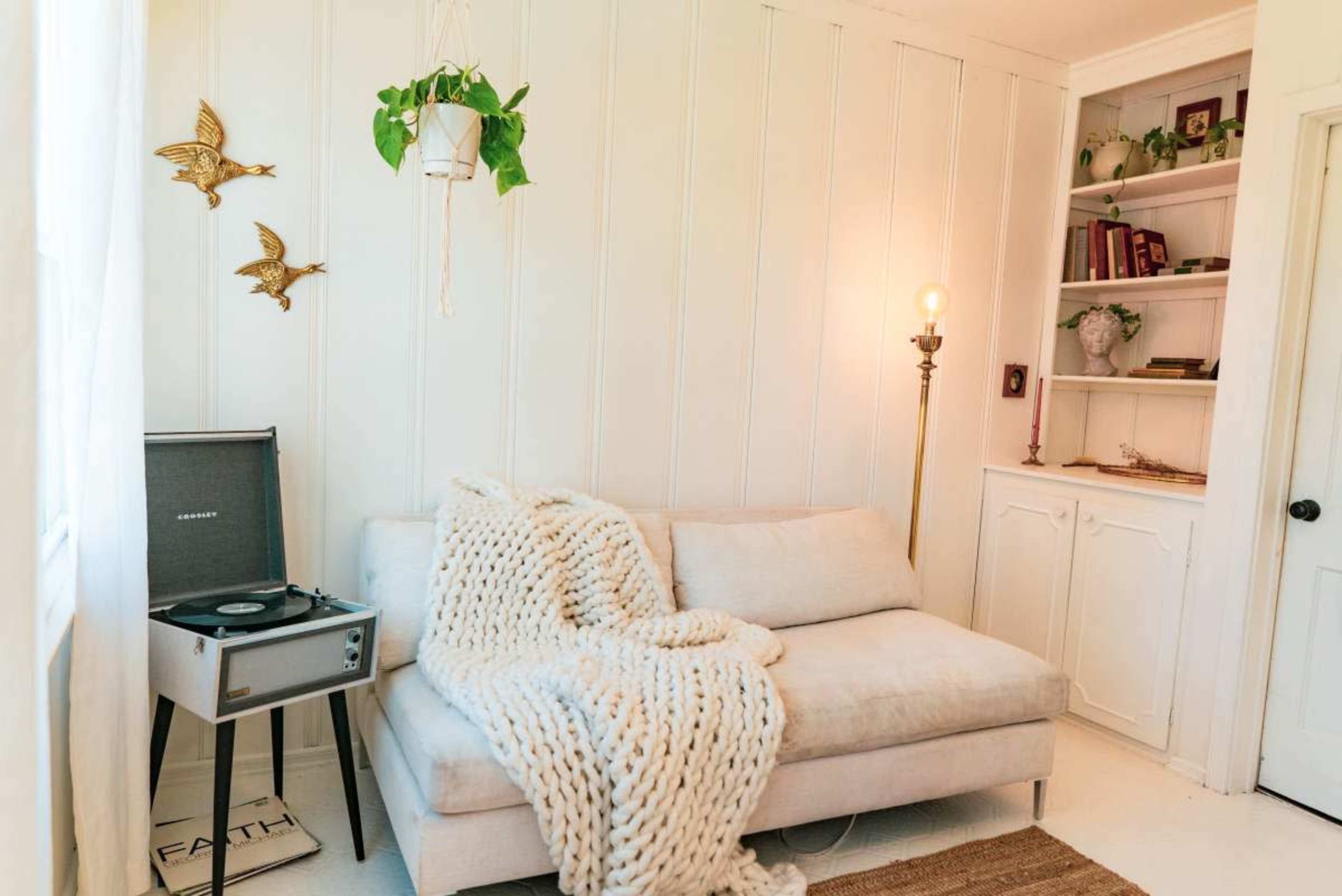 The image shows a cozy room featuring a light-colored sectional sofa draped with a chunky knit blanket, a record player on the floor, and shelves filled with books against a white wall.