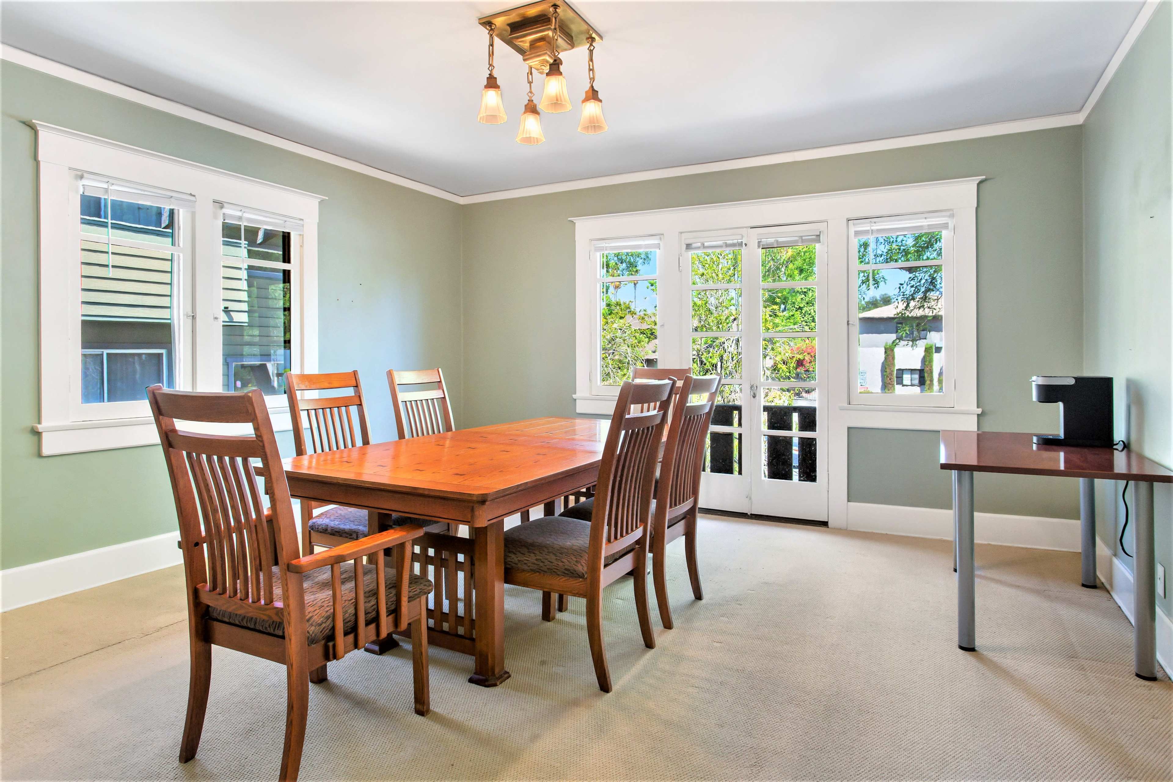 The image shows a spacious dining room featuring a wooden table surrounded by six chairs, with large windows allowing natural light and a side table with a small appliance.