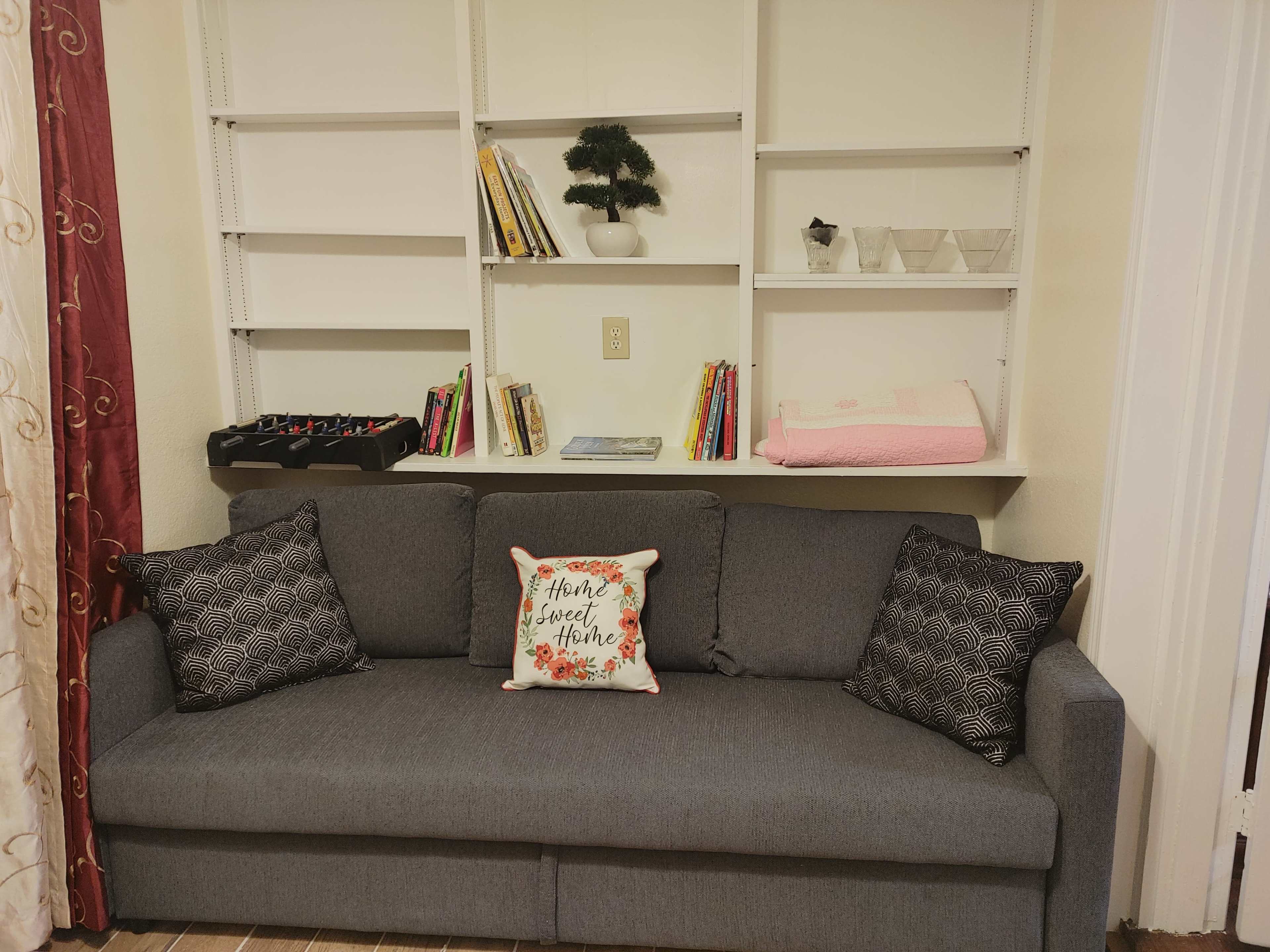 The image shows a gray couch with patterned pillows positioned in front of a wall-mounted shelf displaying books and decorative items.