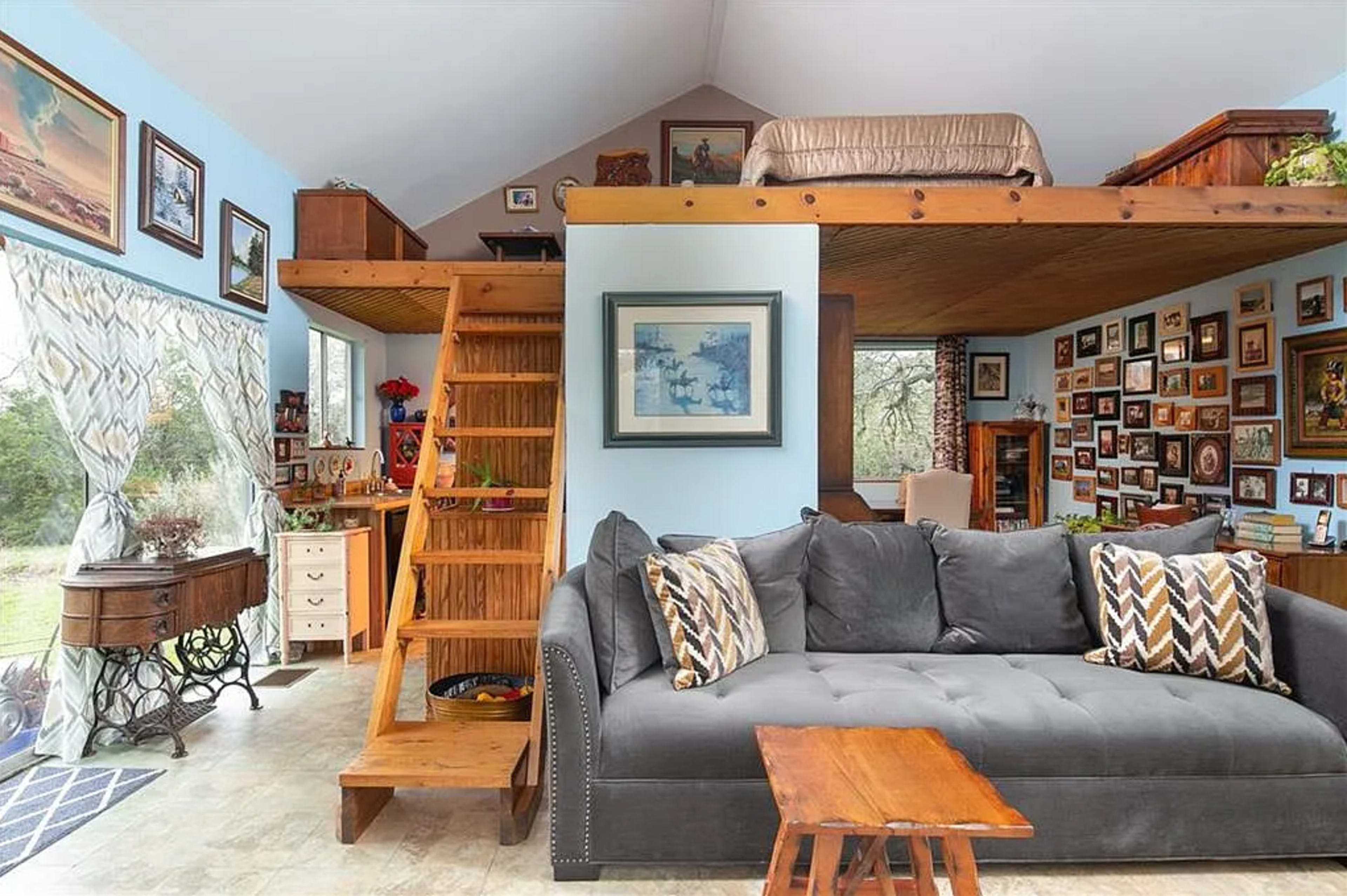 The image depicts a cozy living space featuring a gray couch with patterned pillows, a wooden staircase leading to a lofted area, and walls adorned with various framed pictures.