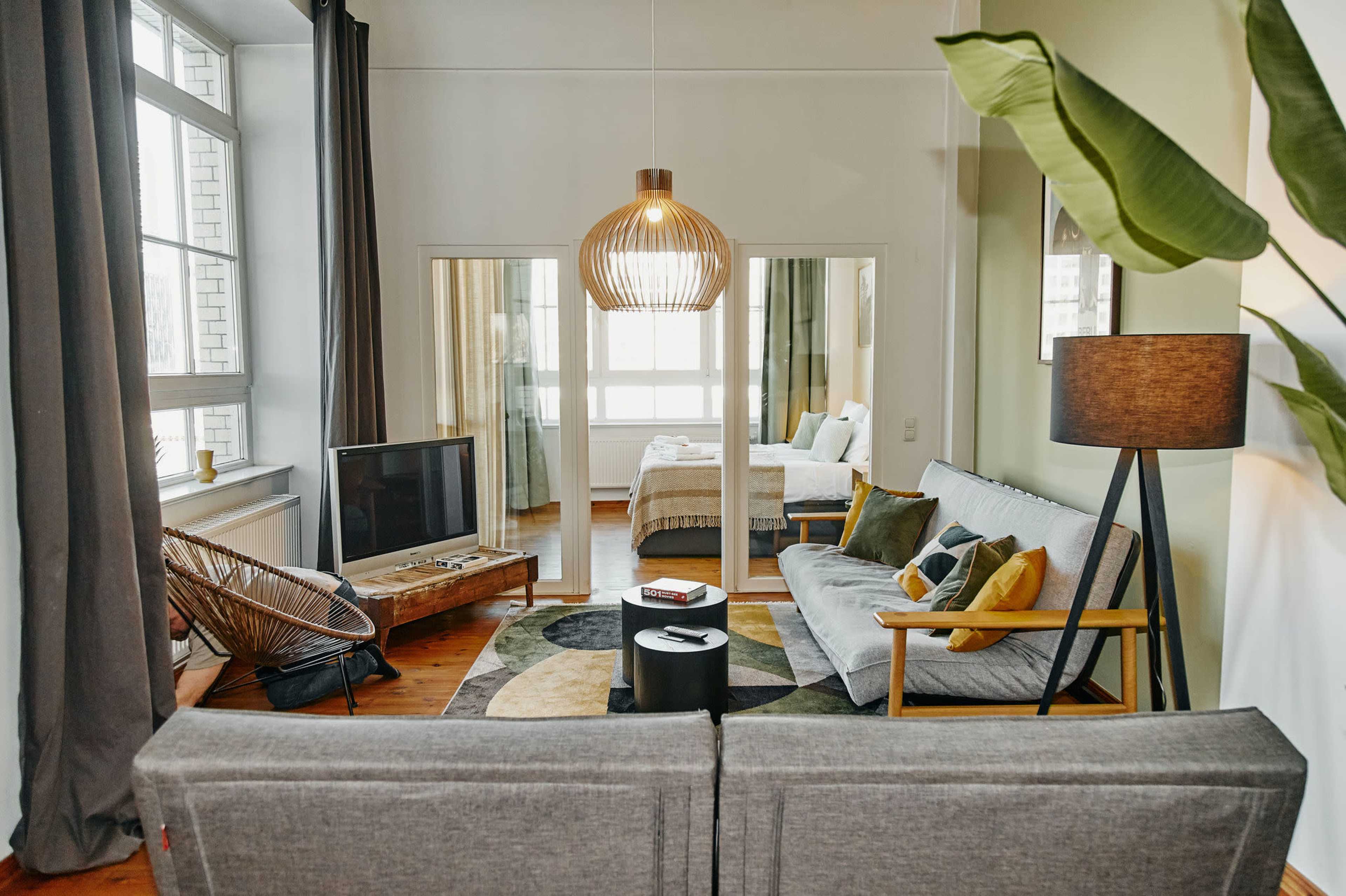 A stylish living space features a gray sofa, a round coffee table, and a television, with a bedroom visible through glass doors in the background.