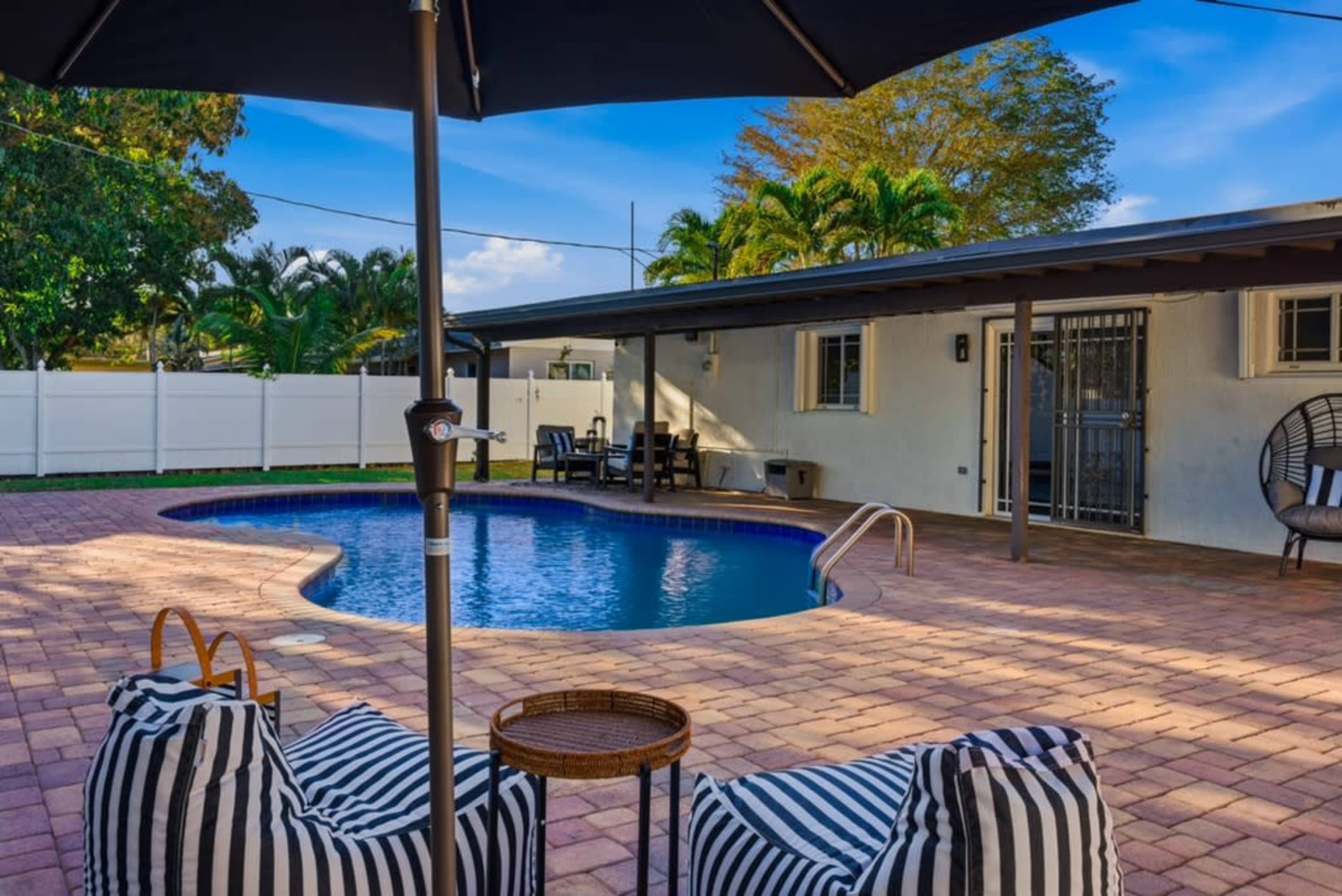The image shows a backyard with a swimming pool surrounded by seating and a shaded area under an umbrella.