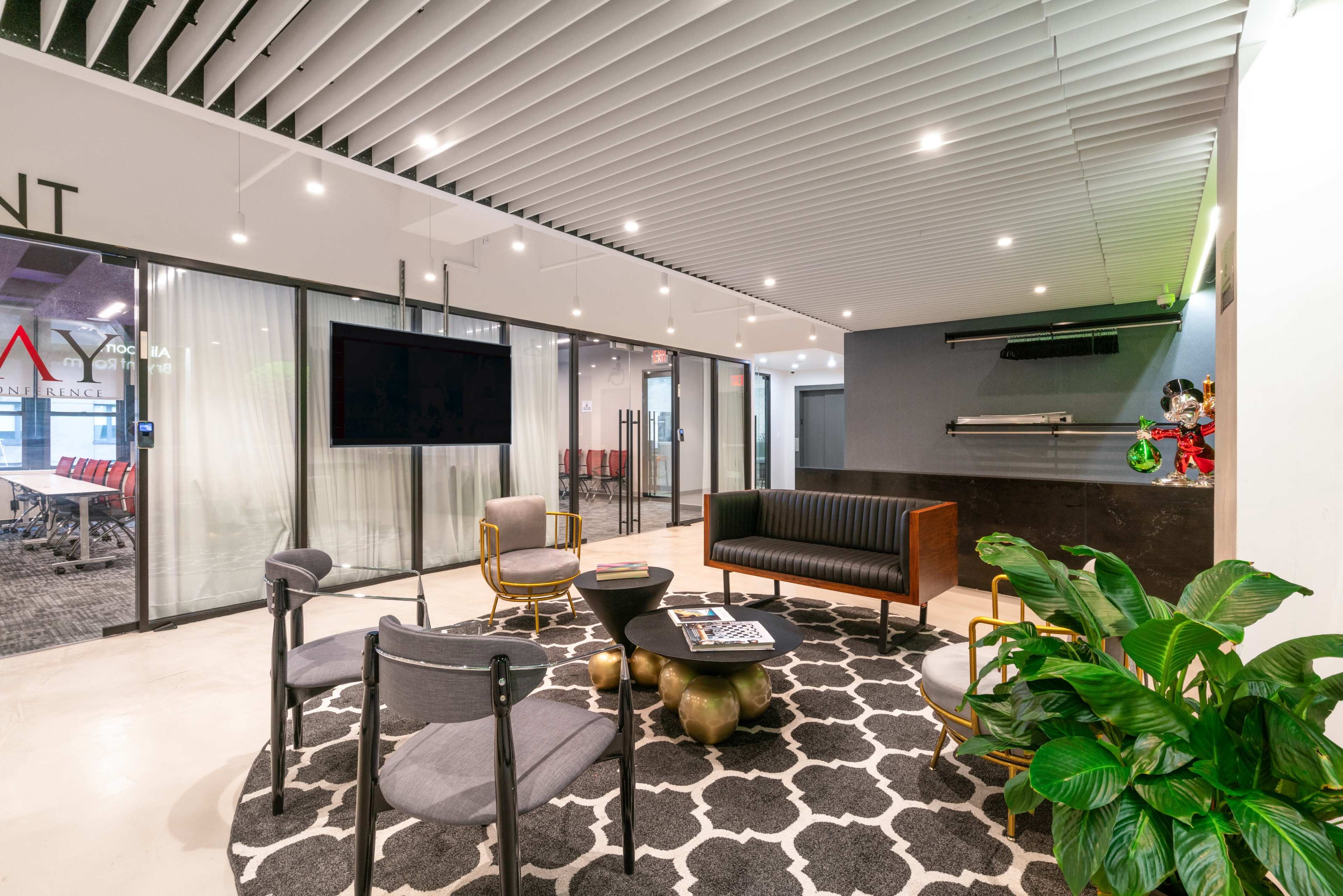 The image shows a modern lobby area with a black leather sofa, gray chairs, a round coffee table, and large plants, set beneath a ceiling with linear lights and acoustic panels.