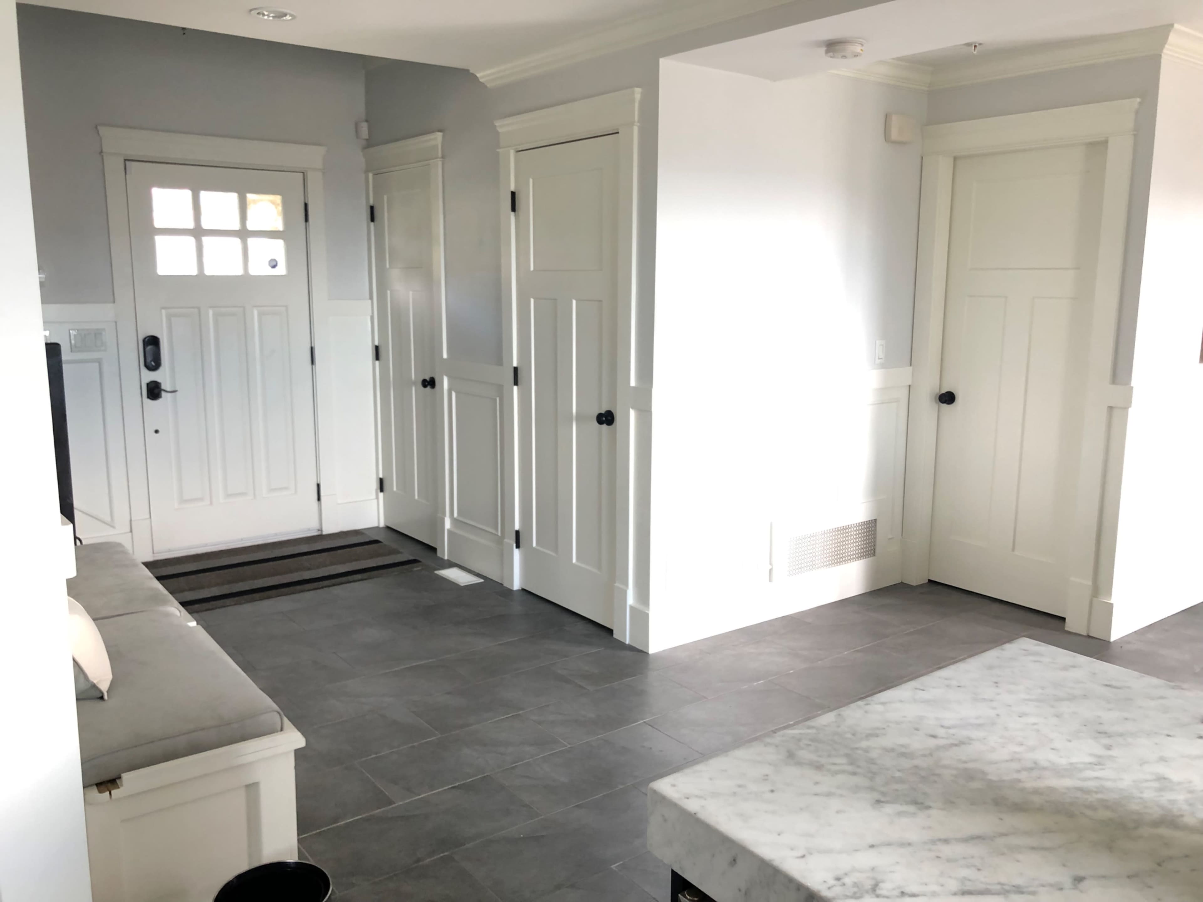 A hallway with multiple closed doors and a front entrance, featuring a gray tiled floor and a white marble countertop in the foreground.