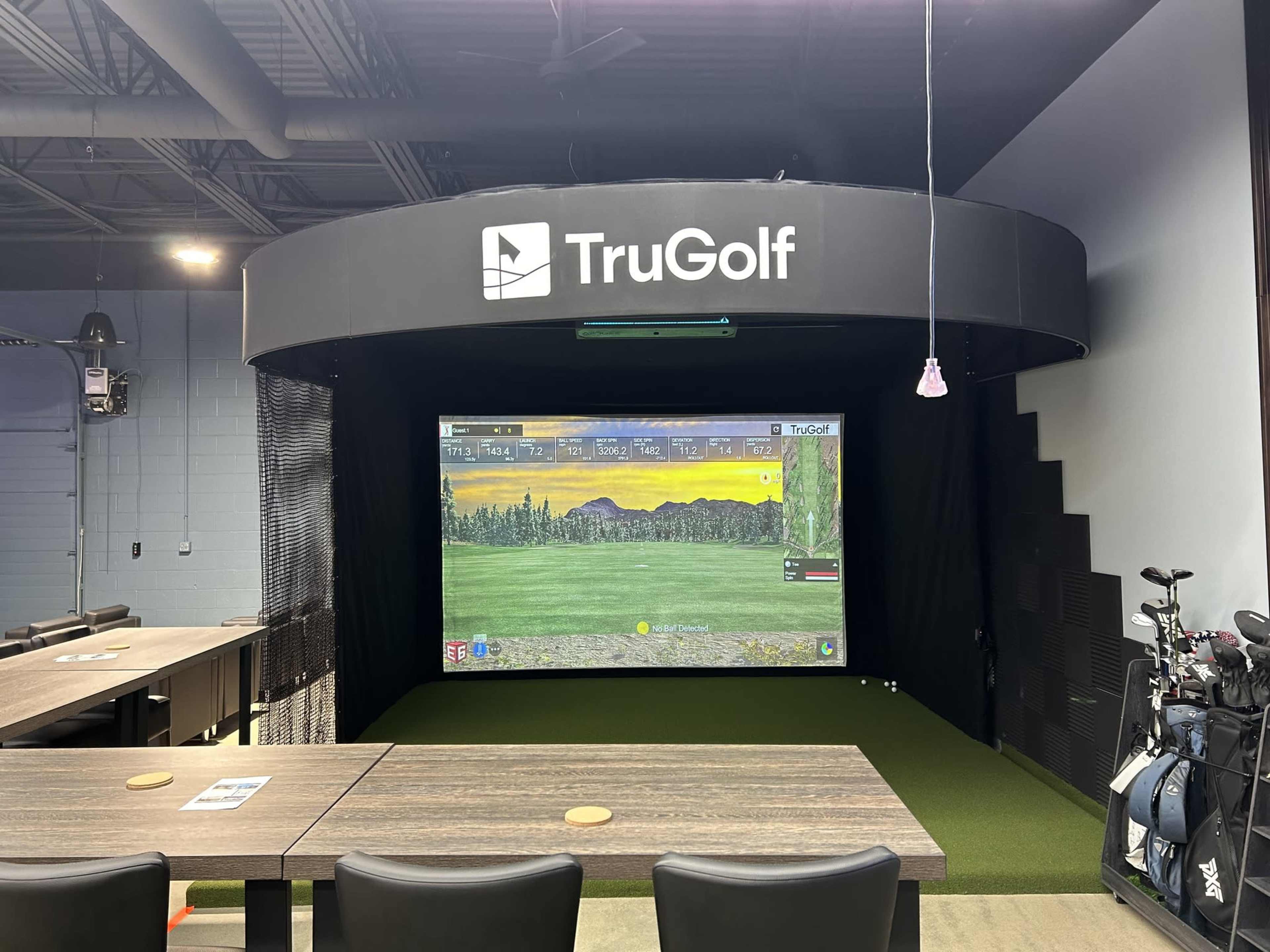 An indoor golf simulator setup featuring a large projection screen, artificial turf, and golf clubs on display nearby.