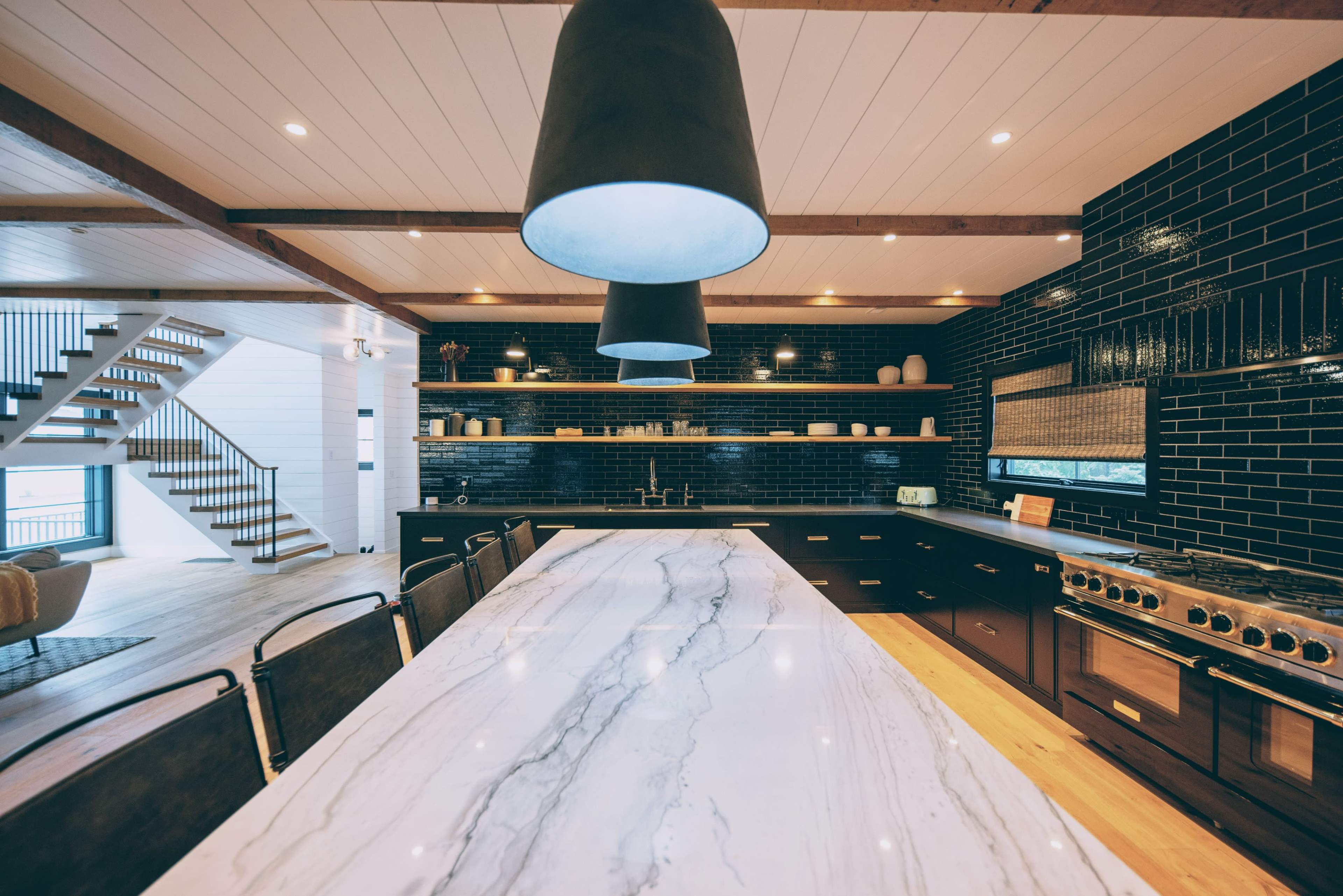 A modern kitchen with a large marble countertop, pendant lights, dark cabinetry, and a staircase in the background.