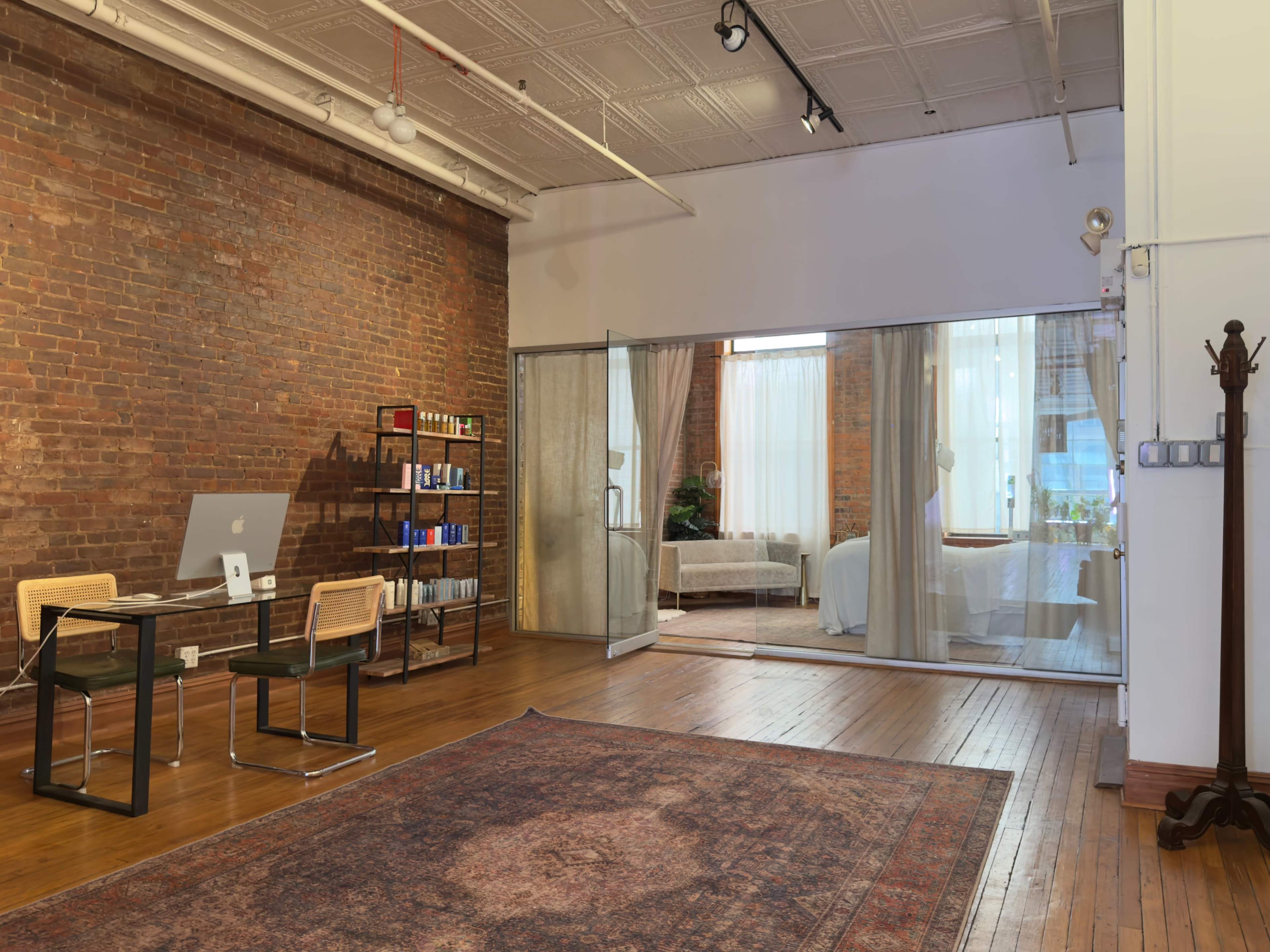 The image shows an industrial-style room with exposed brick walls, a wooden floor, a glass partition leading to a bedroom area, and a desk with a computer setup.