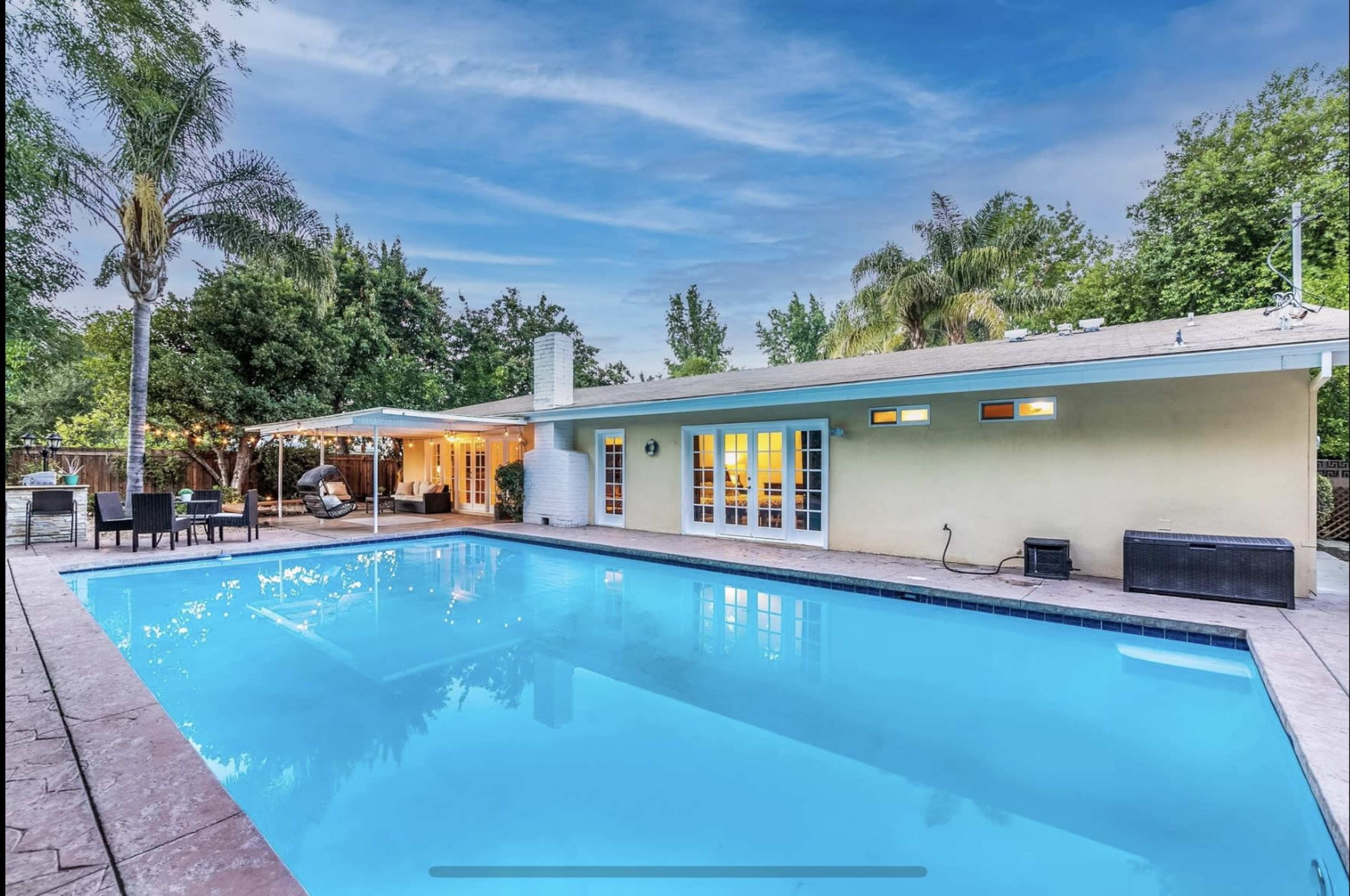 A backyard features a clear pool surrounded by a patio, with a covered seating area and a light-colored house in the background.