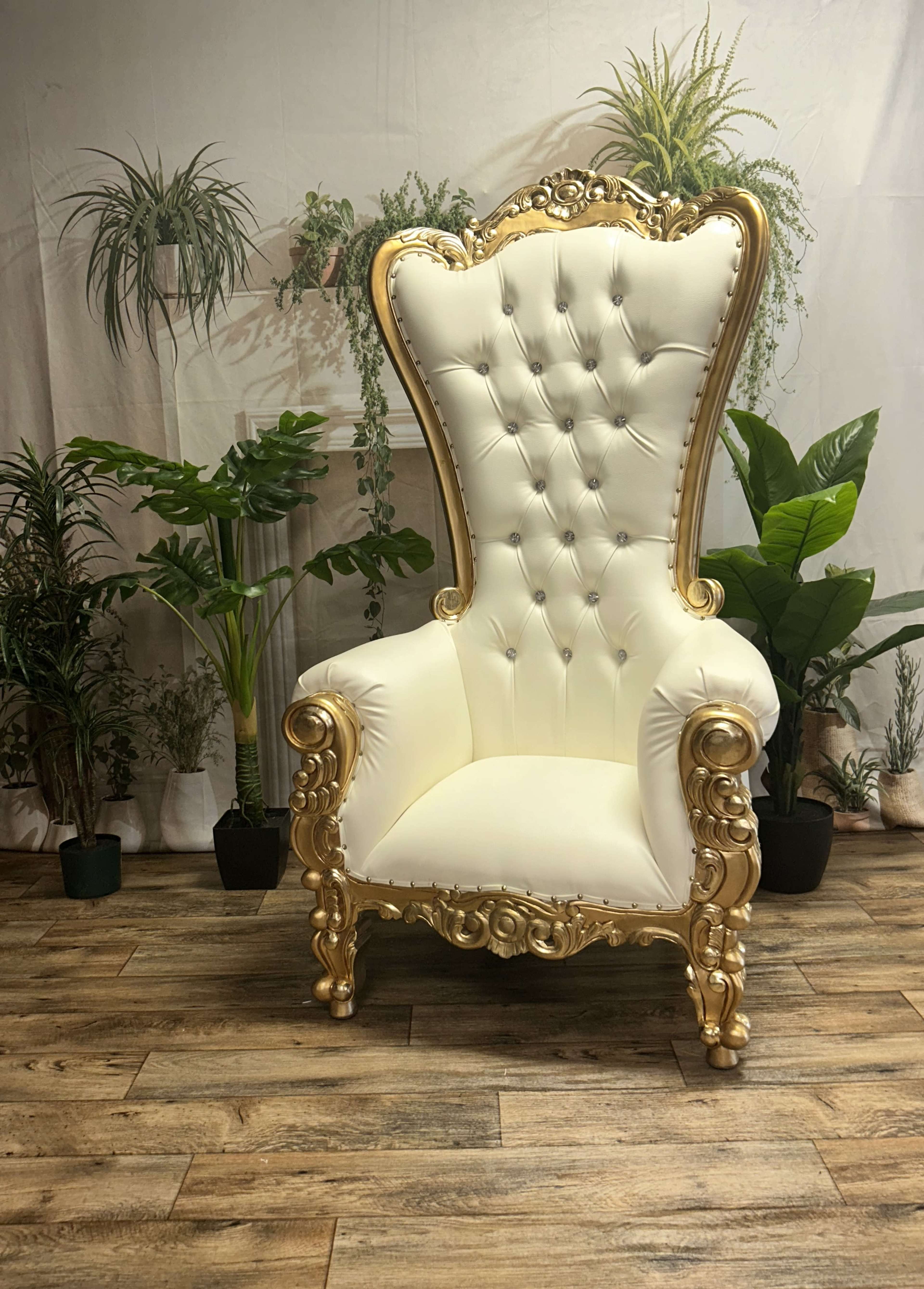 A white and gold ornate chair with a high back is positioned against a backdrop of indoor plants.