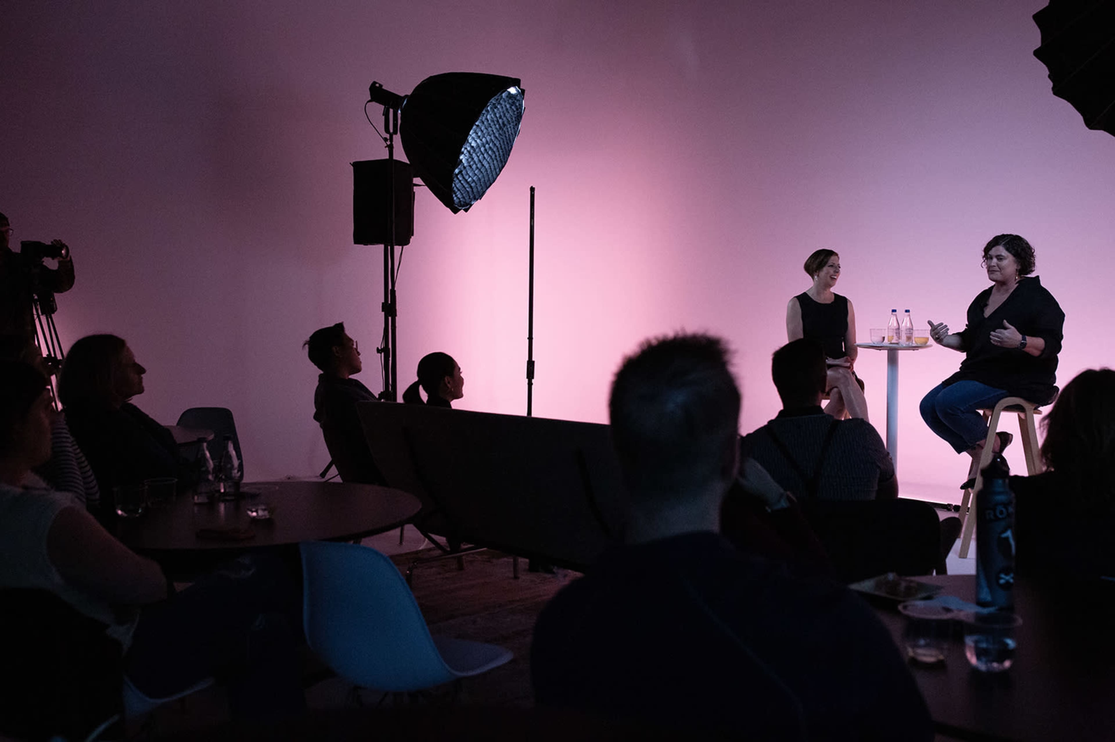 A panel discussion takes place on a stage with two speakers seated and an audience observing in a softly lit room.