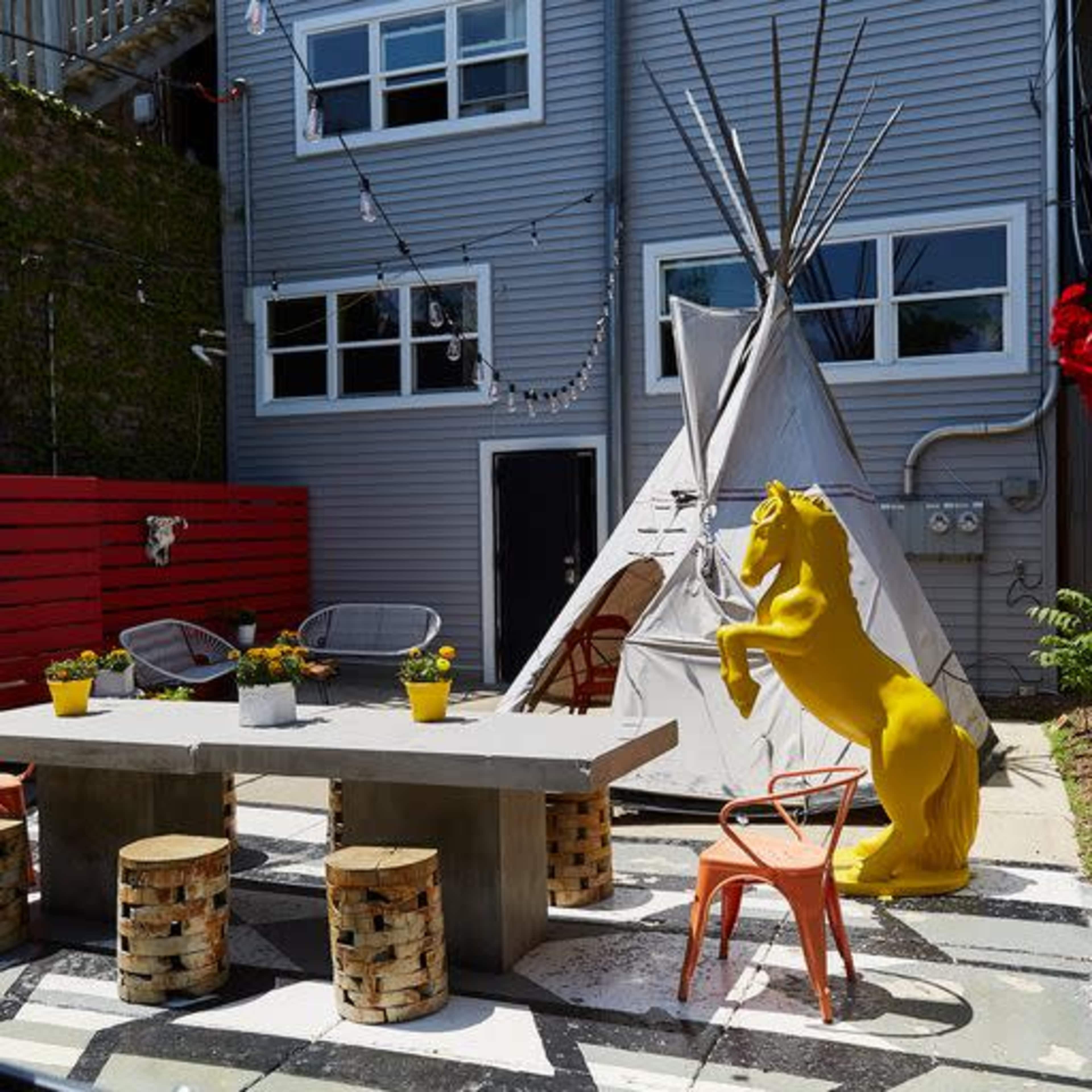 The image shows a stylish backyard featuring a table surrounded by stools, a teepee, and a yellow horse sculpture against a backdrop of wooden paneling and modern windows.