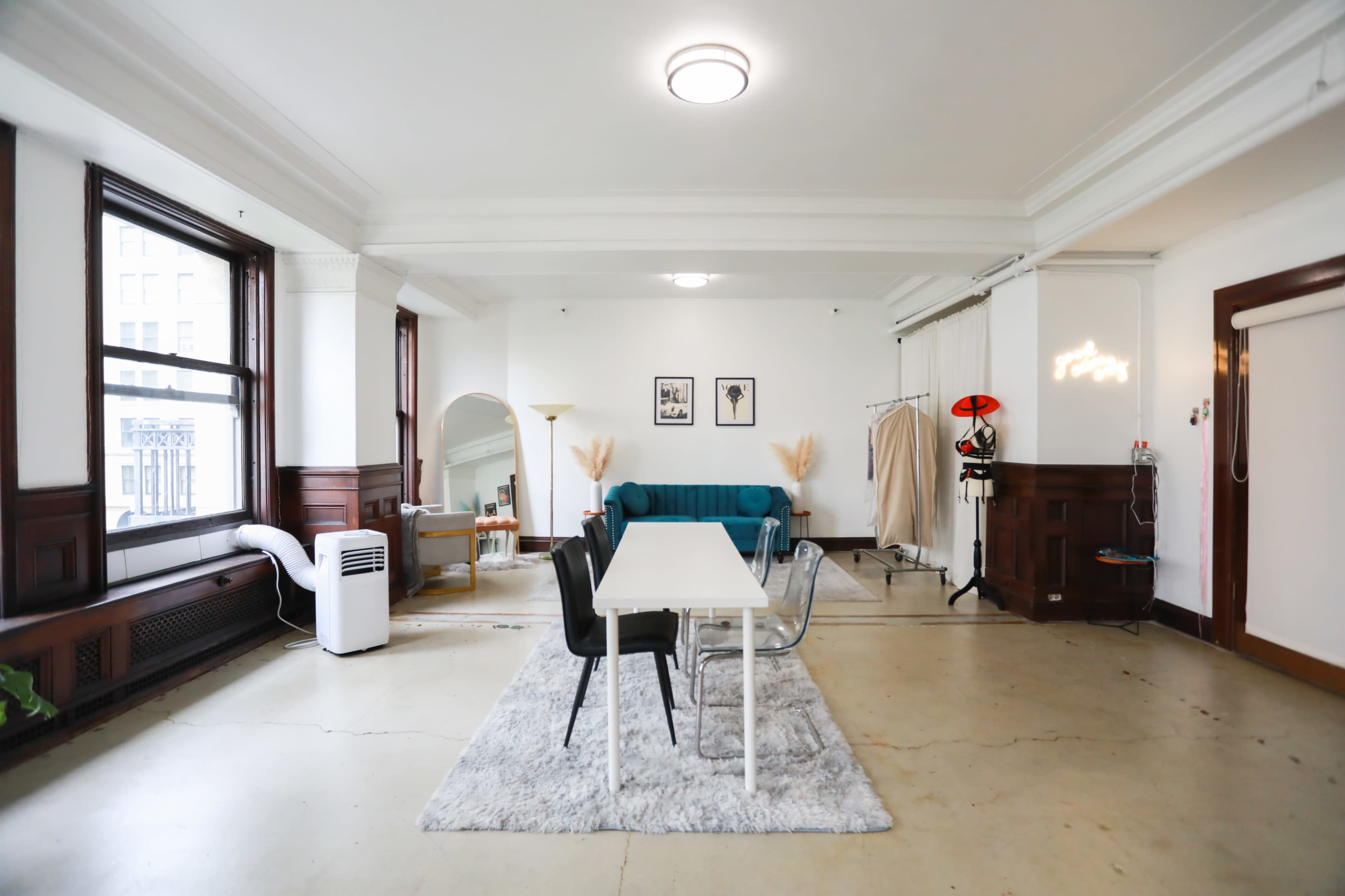 A spacious, minimalistic living area featuring a dining table surrounded by chairs, a teal sofa, and a clothing rack in an airy room with large windows.