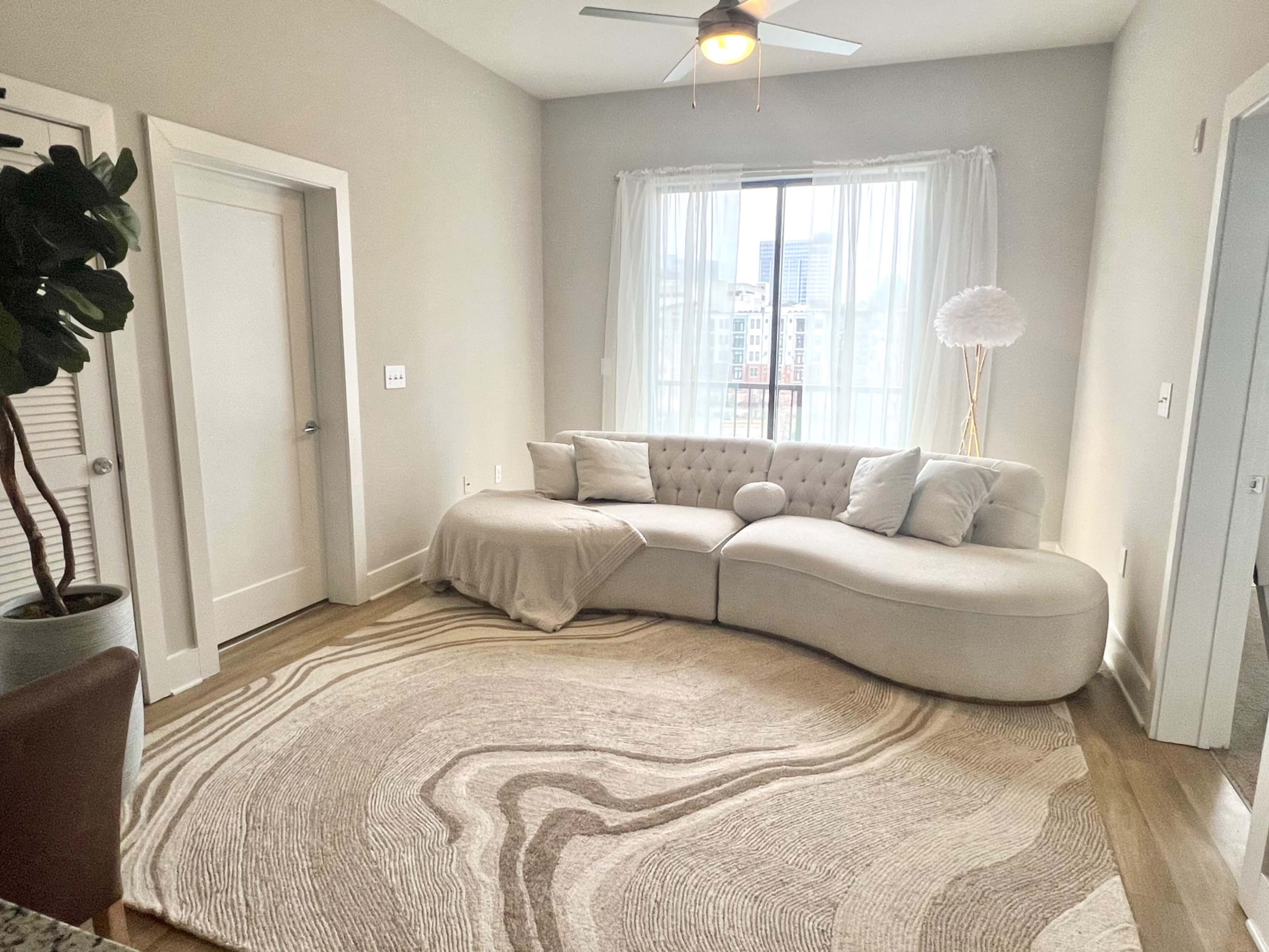A modern living room featuring a curved white sectional sofa, a round area rug with a wave design, and large windows dressed with sheer curtains.