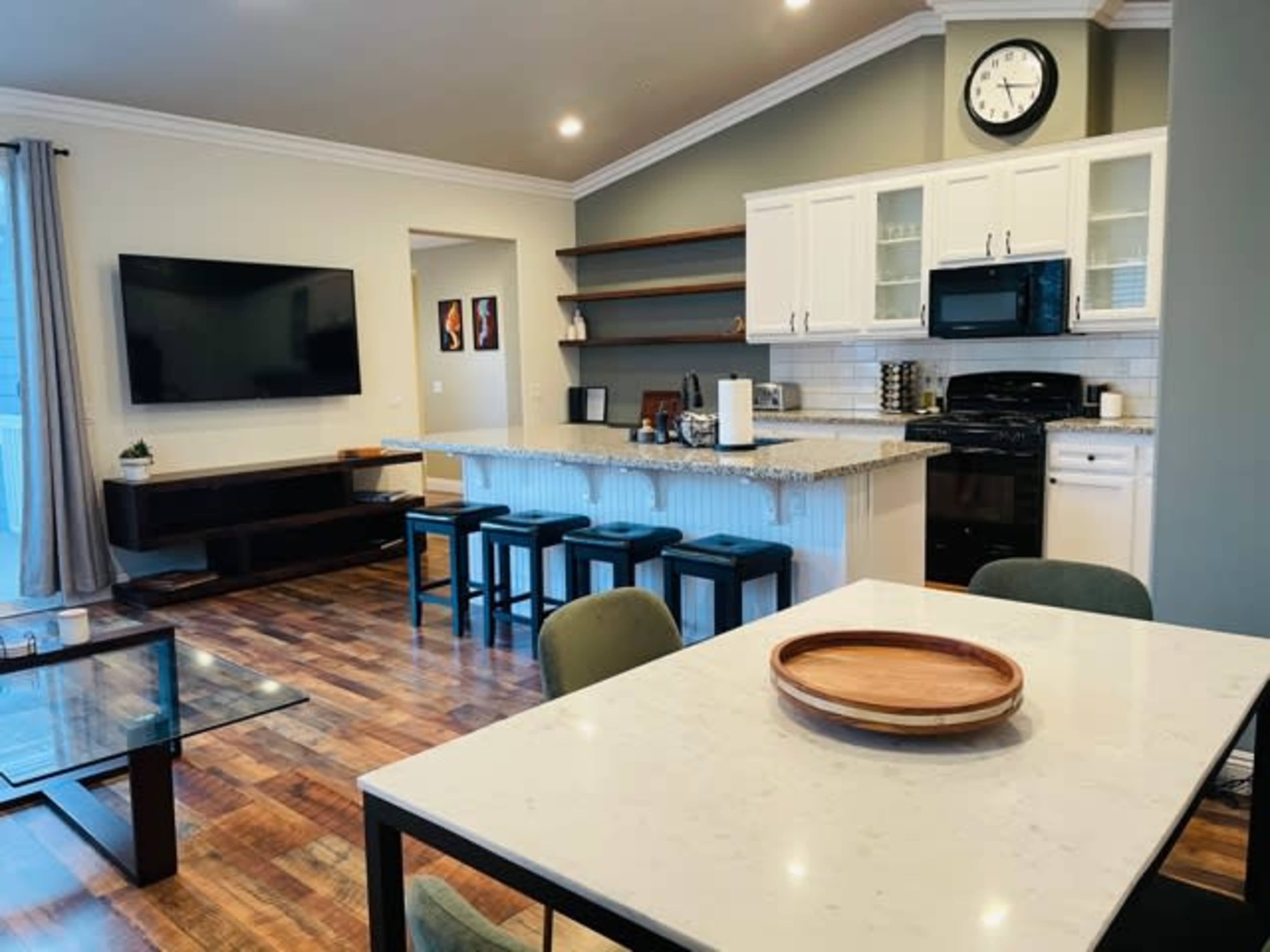 The image shows a modern kitchen and living area featuring a granite island with bar stools, a television mounted on the wall, and a dining table with a tray on it.