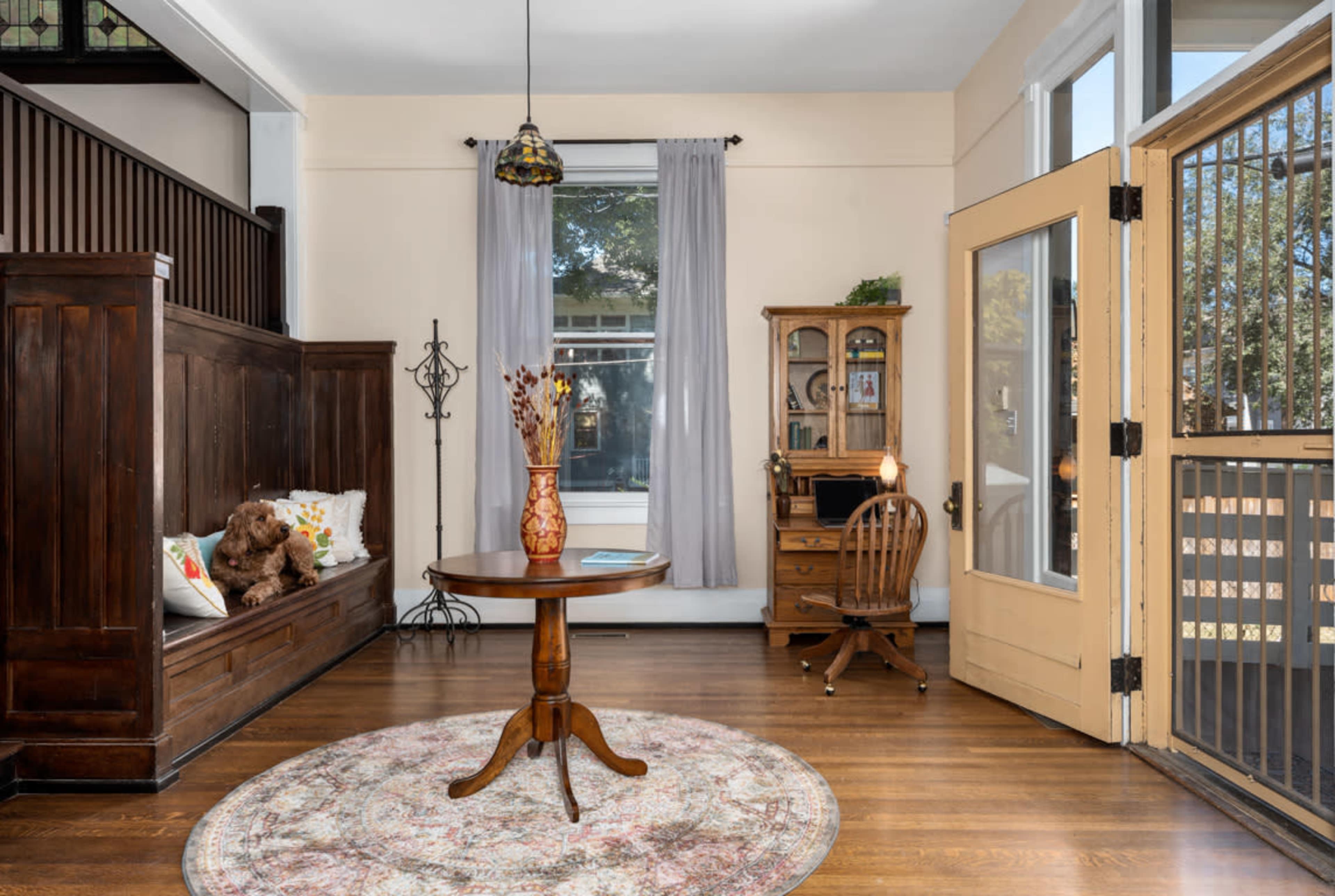 A well-lit entryway features a round wooden table, a vintage writing desk, and a cozy nook with a dog on a cushioned seat.