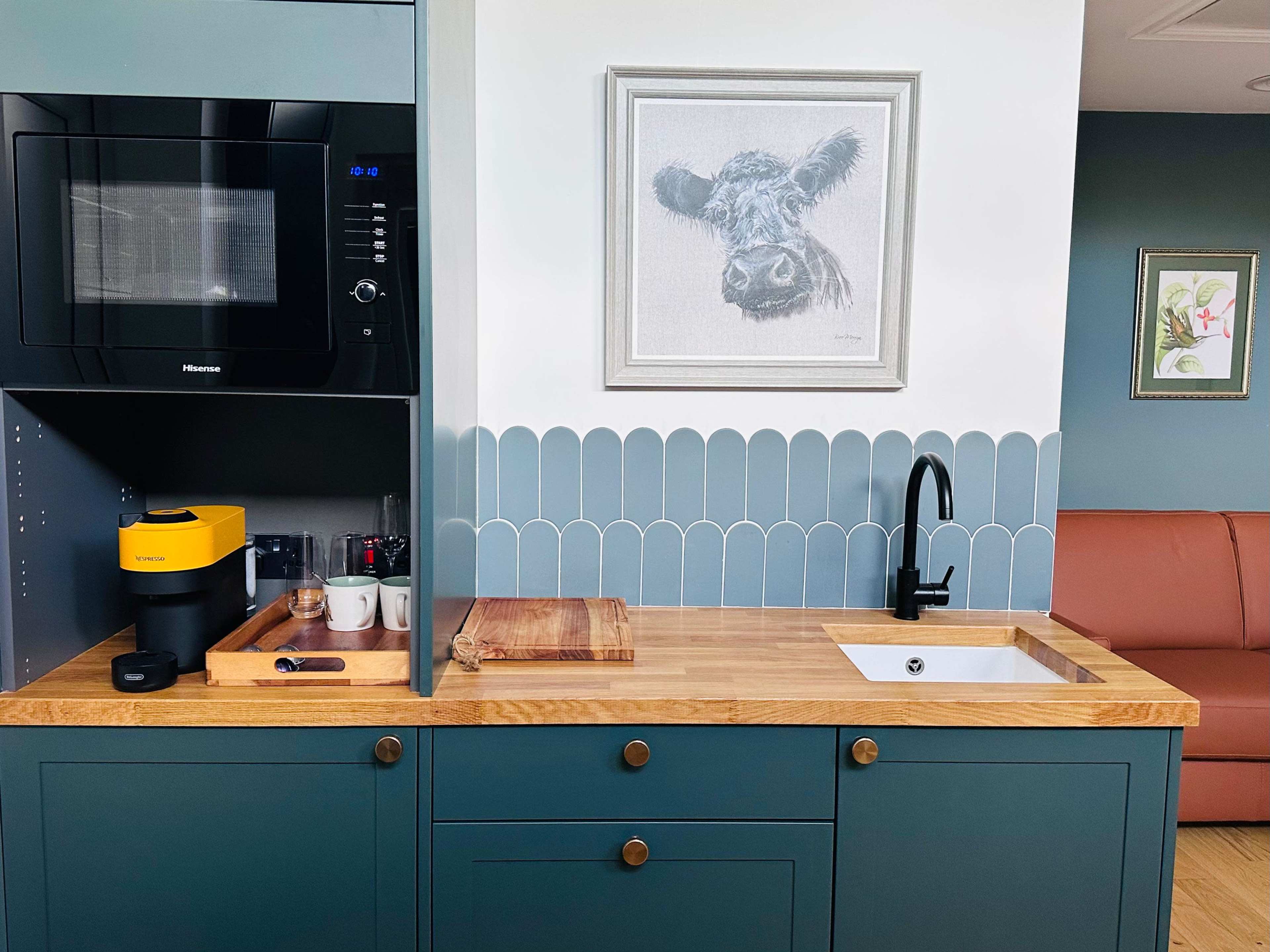 The image shows a modern kitchen area featuring a microwave, a coffee maker, a wooden cutting board, and a small sink with a black faucet, all set against a backdrop of blue cabinetry and tiled wall decor.