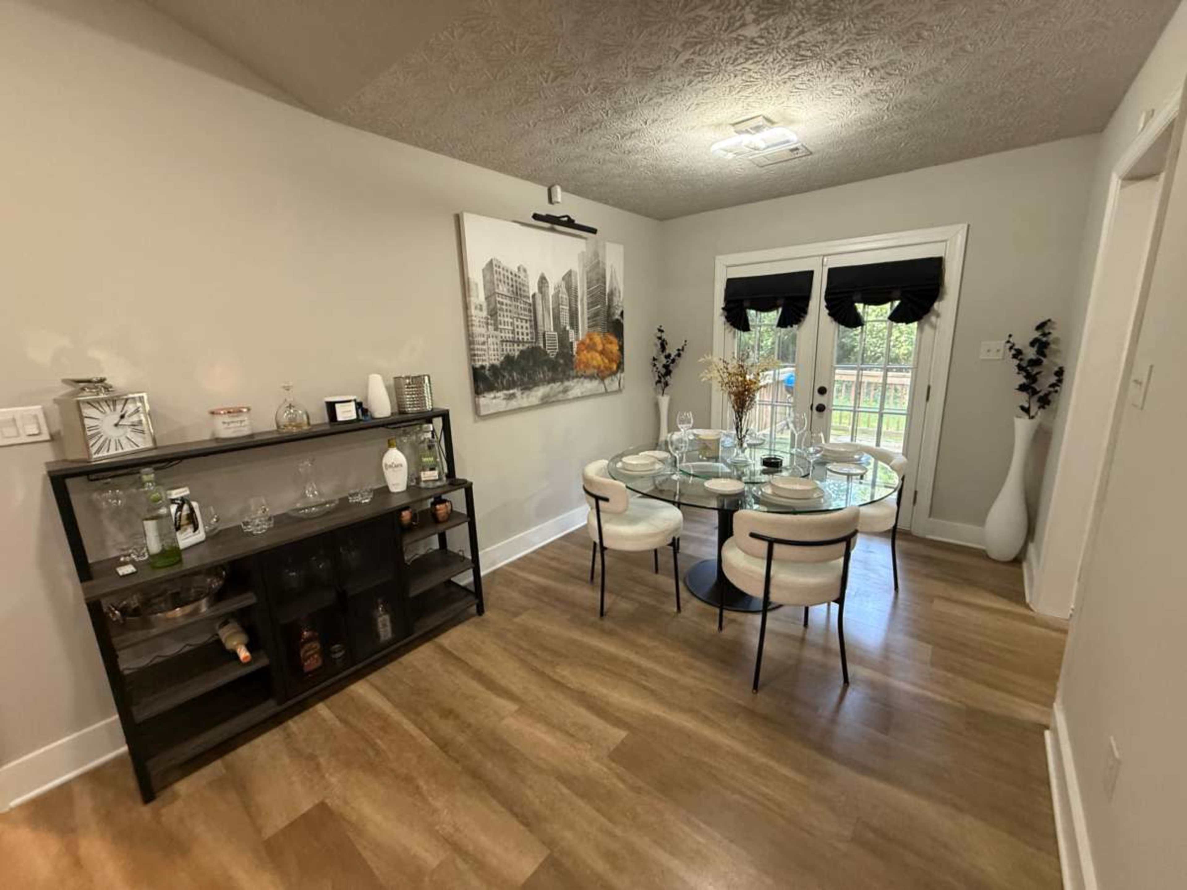 The image shows a dining area with a round glass table surrounded by six chairs, a large wall-mounted artwork, and a display cabinet with glassware and decorative items.