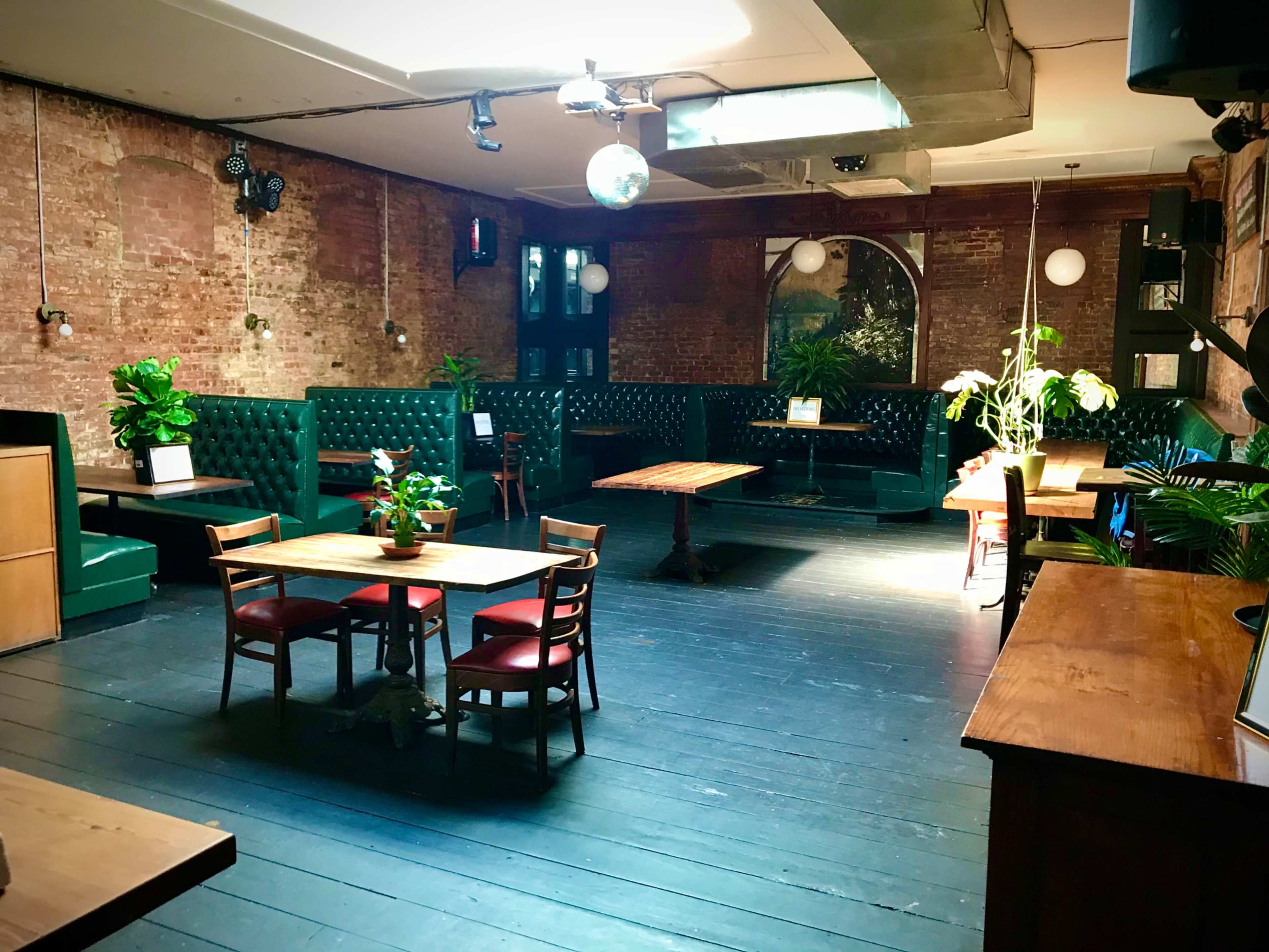 An empty interior of a restaurant or bar with green upholstered seating, wooden tables, and potted plants against exposed brick walls.