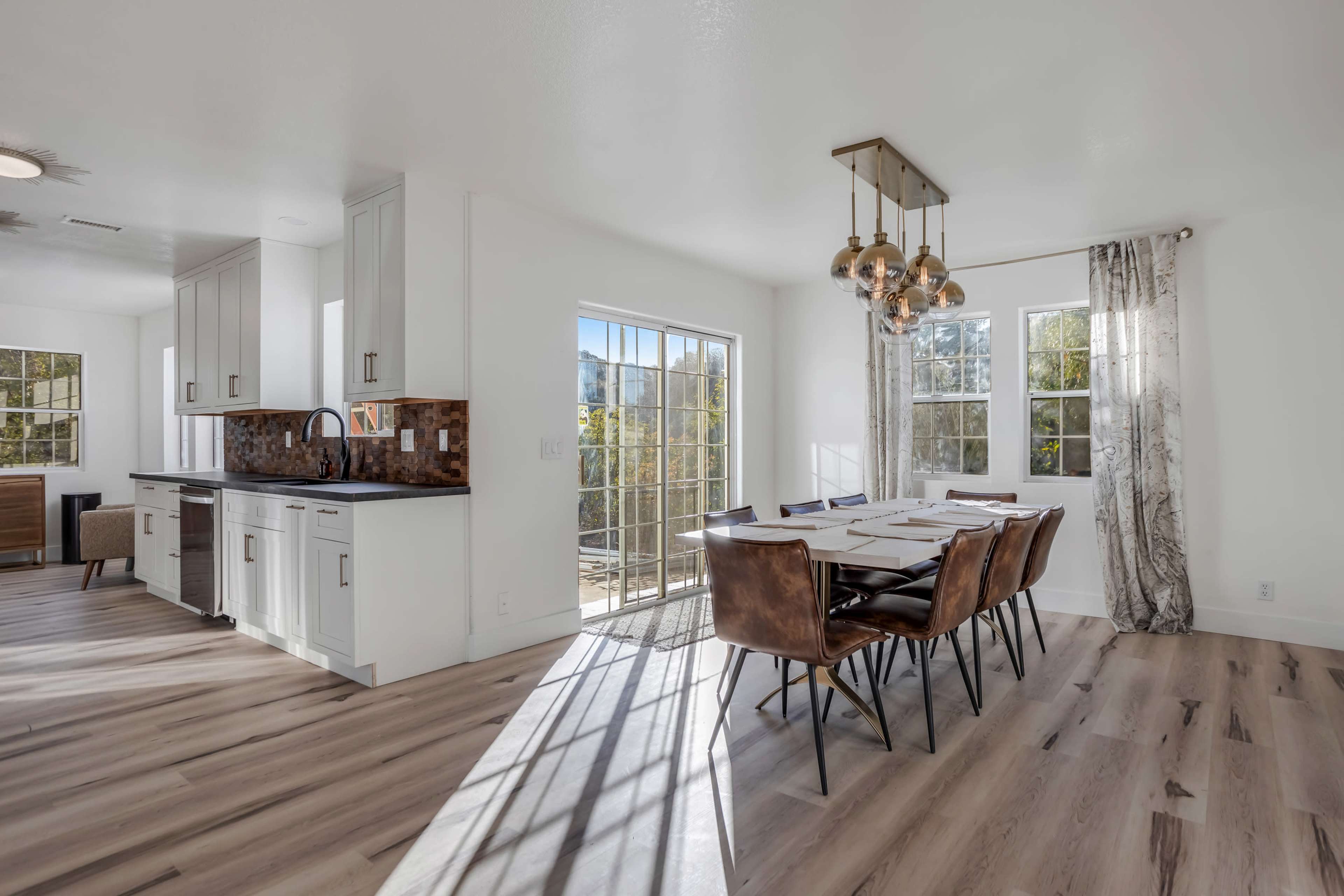 A modern dining area features a large wooden table with brown leather chairs, illuminated by a pendant light, adjacent to a kitchen space with white cabinets and large windows.