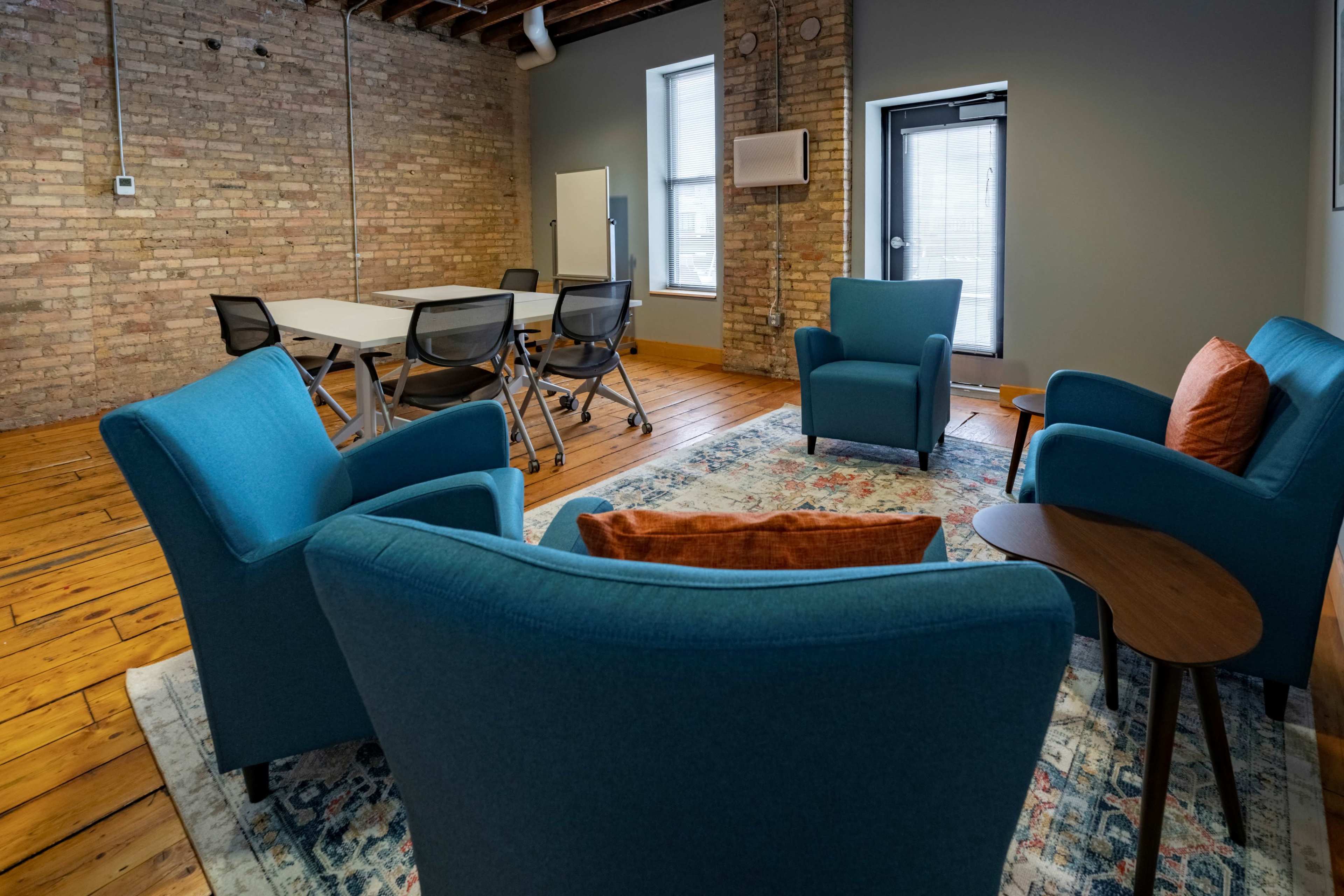 The image shows a conference room with four blue chairs arranged around a small table on a patterned rug, with a large table in the background and exposed brick walls.