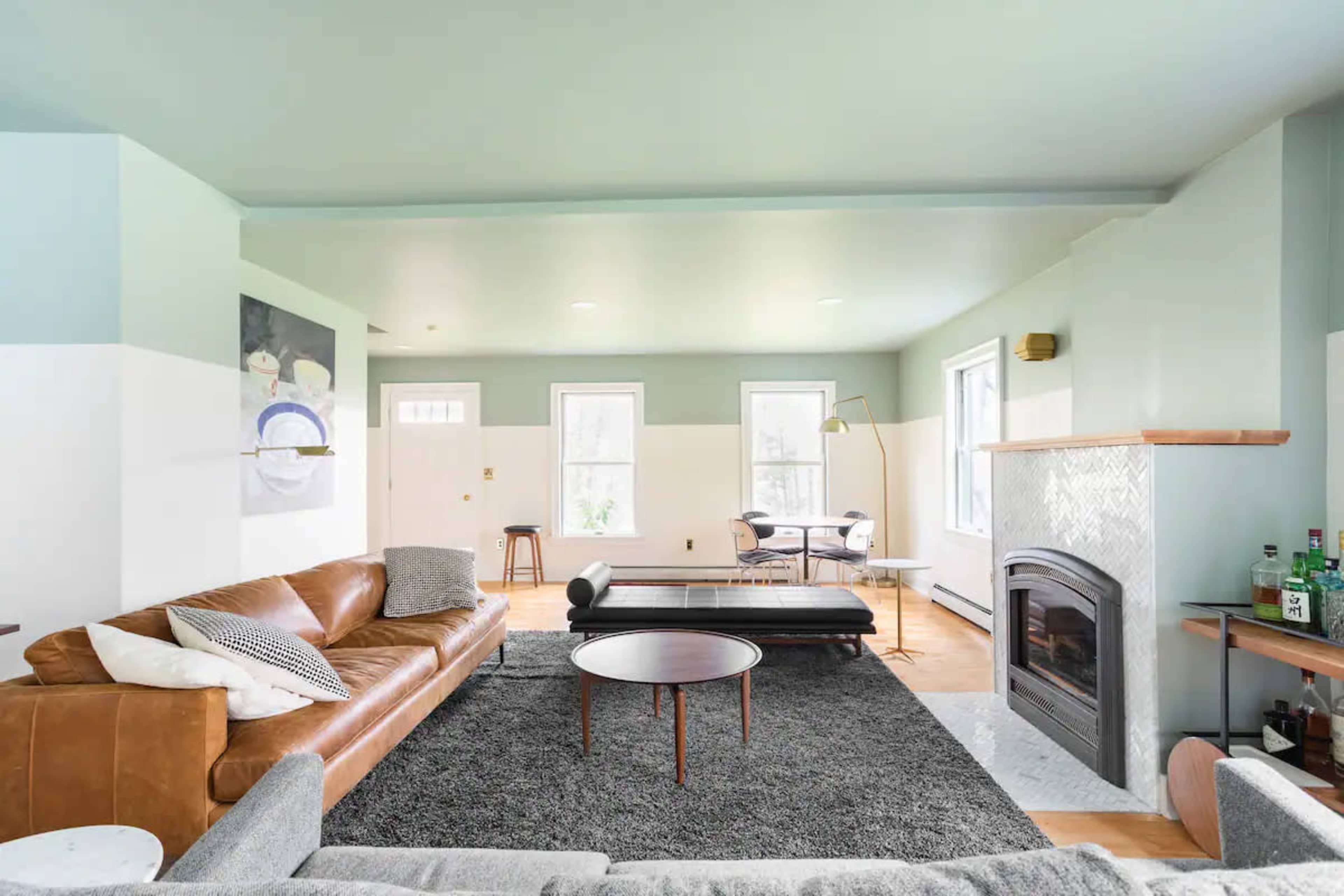 A modern living room features a leather sectional sofa, a round coffee table, a patterned rug, and a fireplace, with large windows allowing natural light.