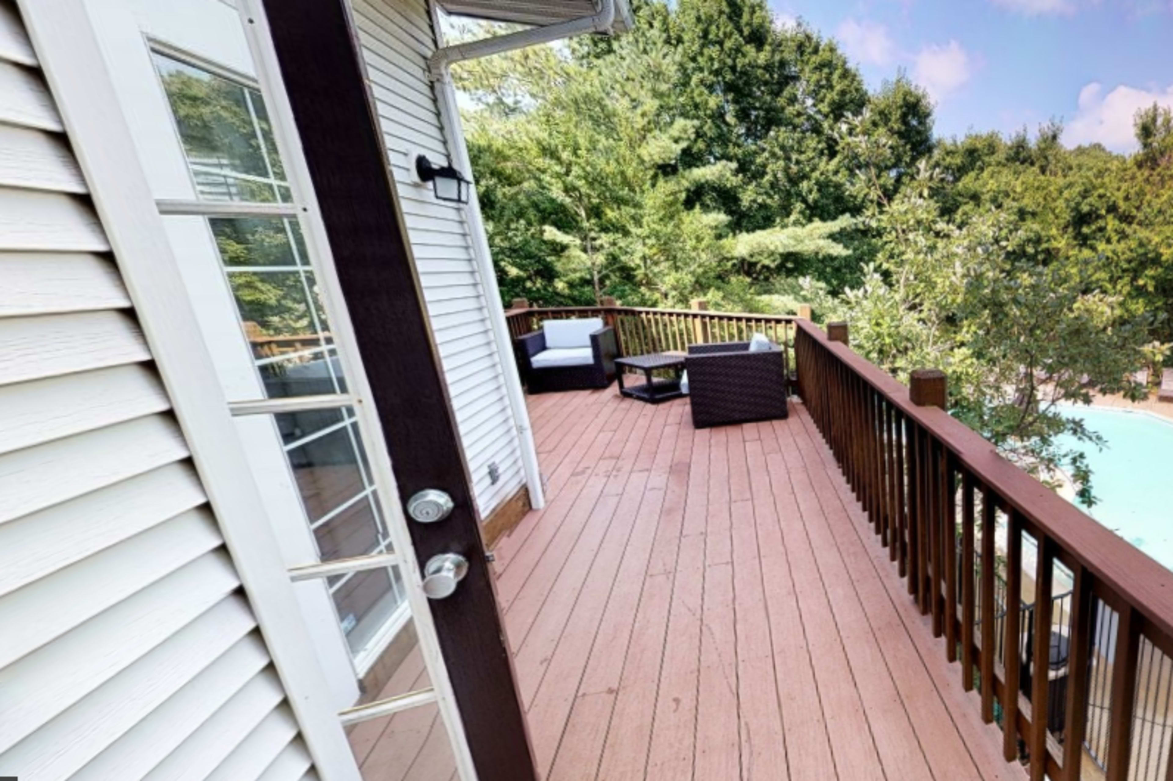 The image shows a wooden deck attached to a house, with seating arrangements and a view of greenery in the background.