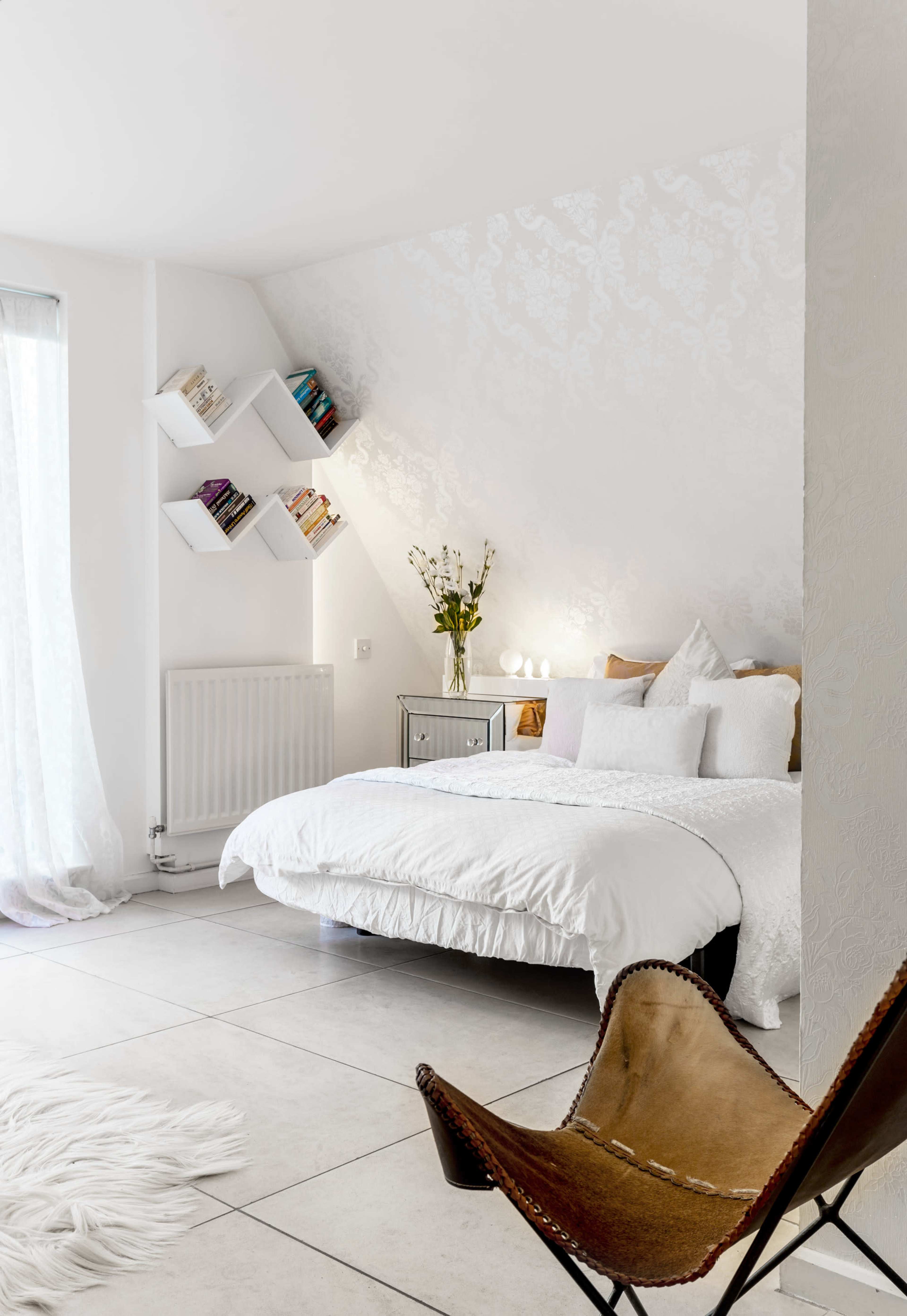 A modern bedroom with a large bed, a decorative chair, and wall shelves holding books, all set against a light-colored backdrop.