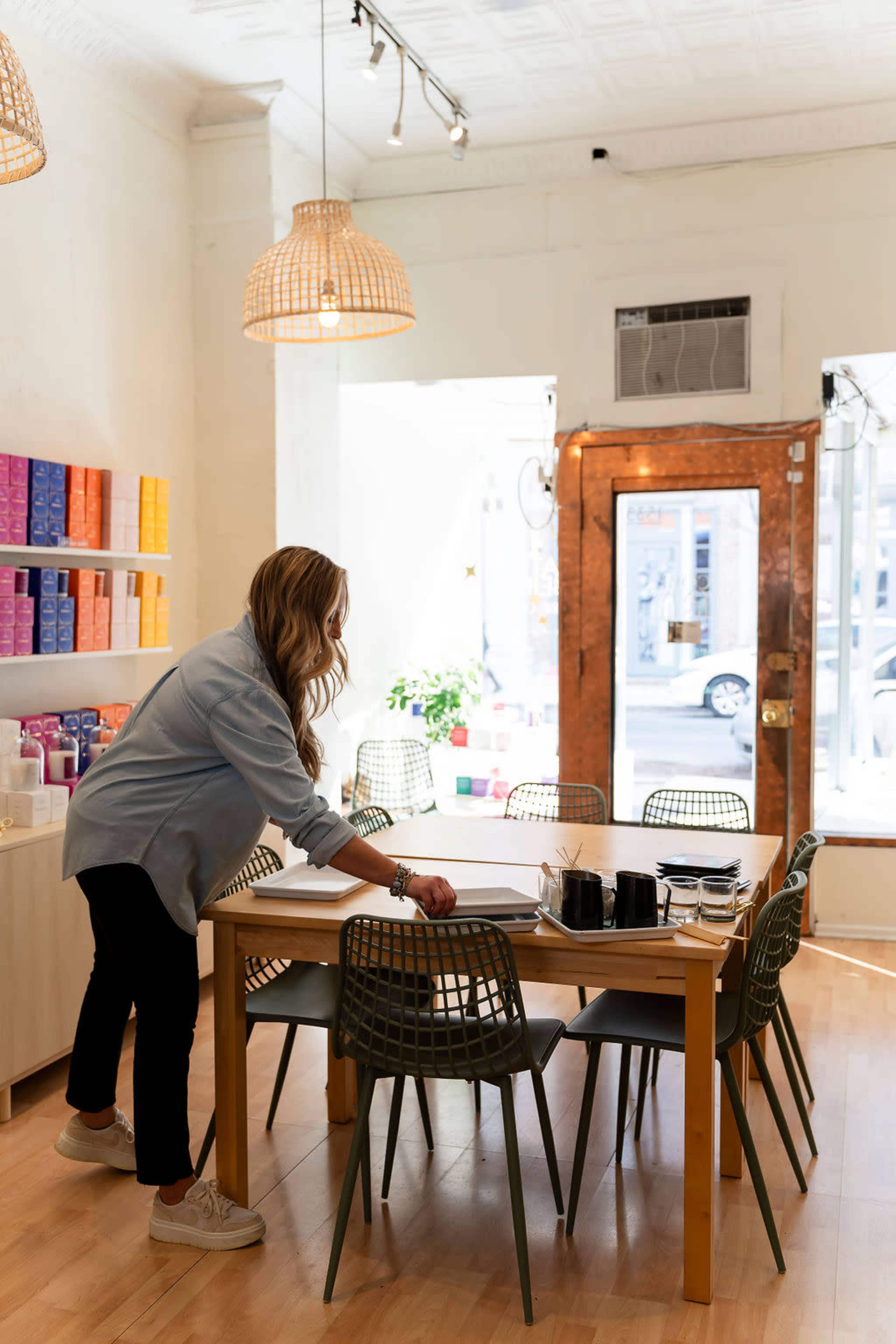 A woman adjusts items on a dining table in a brightly lit room with modern decor and a large window.