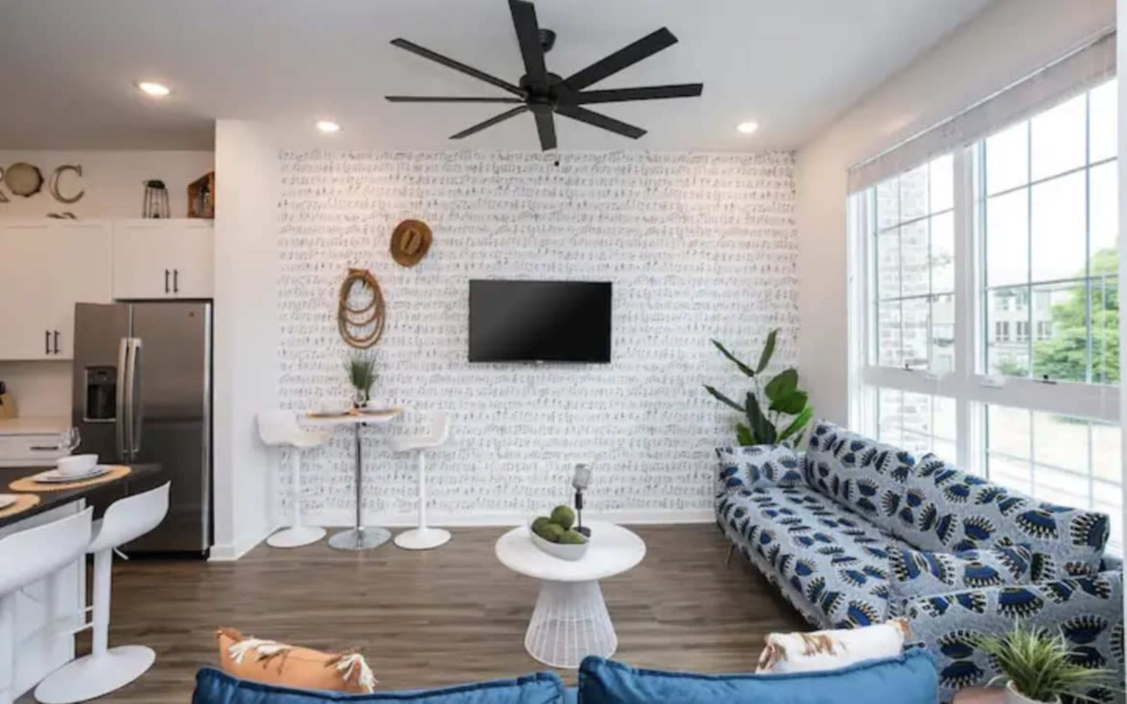 A modern living space features a patterned sofa, a wall-mounted television, and a dining area with bar stools, complemented by large windows and decor elements.