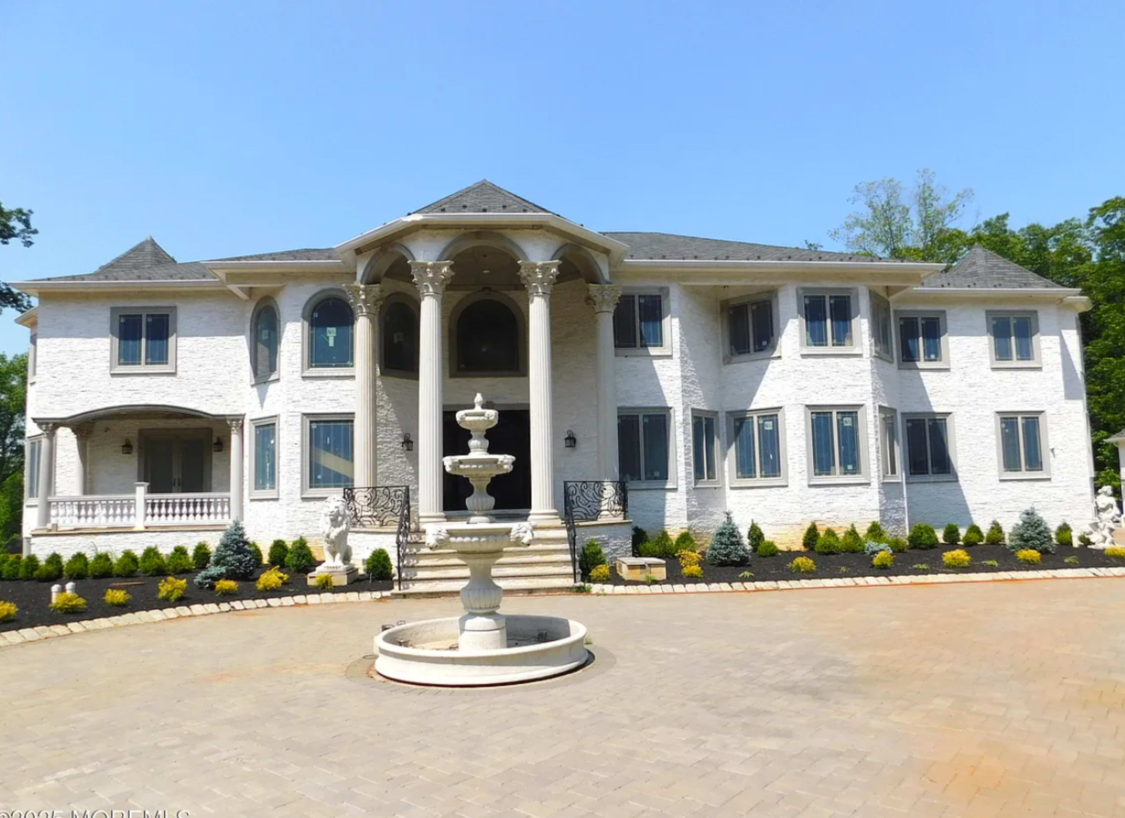 A large, multi-story mansion features a central entrance with four columns and a circular fountain in the driveway.