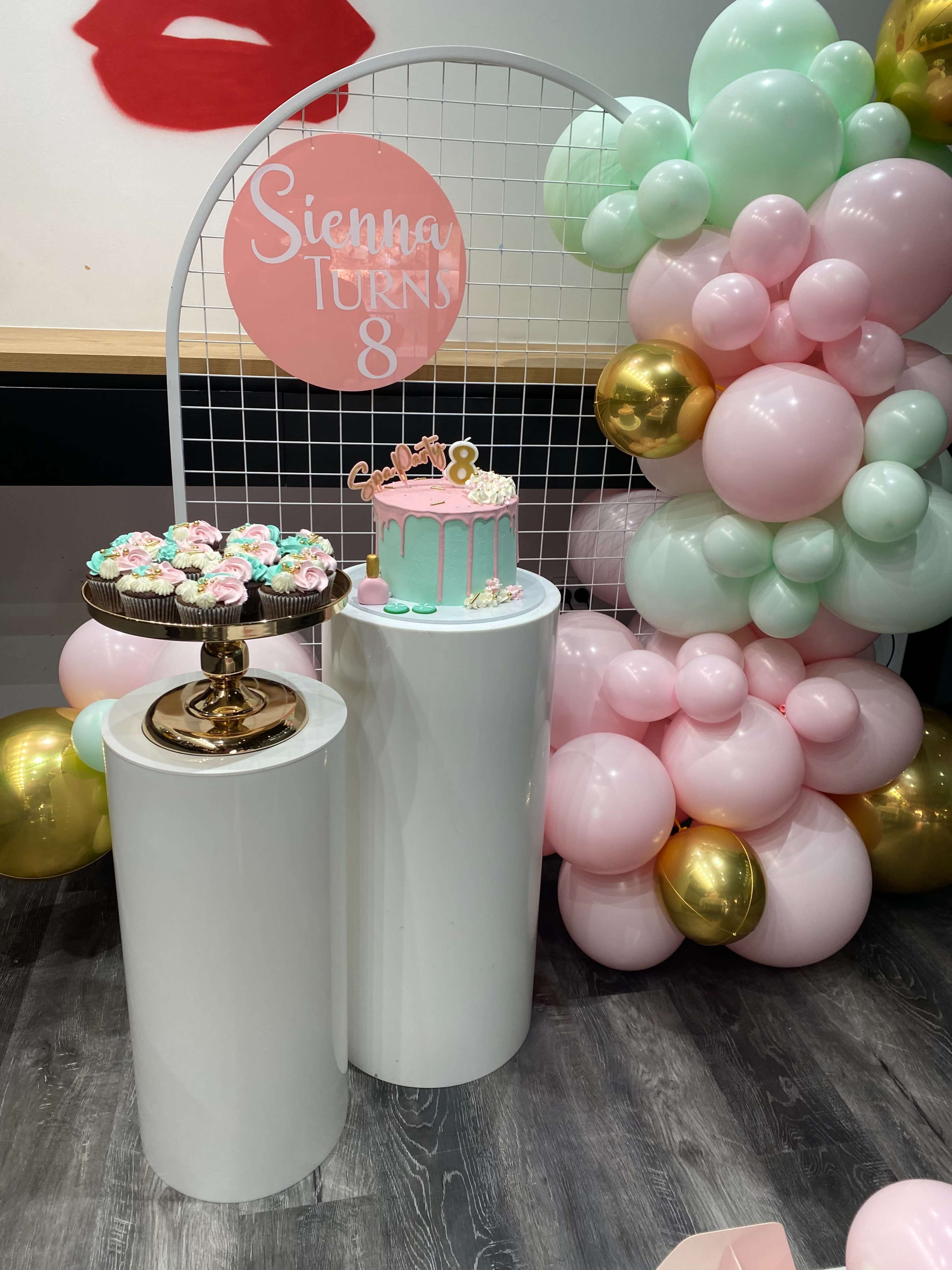 A birthday celebration setup with a pink and mint green balloon backdrop, a cake on a pedestal labeled "Sienna Turns 8," and a decorative stand of cupcakes.