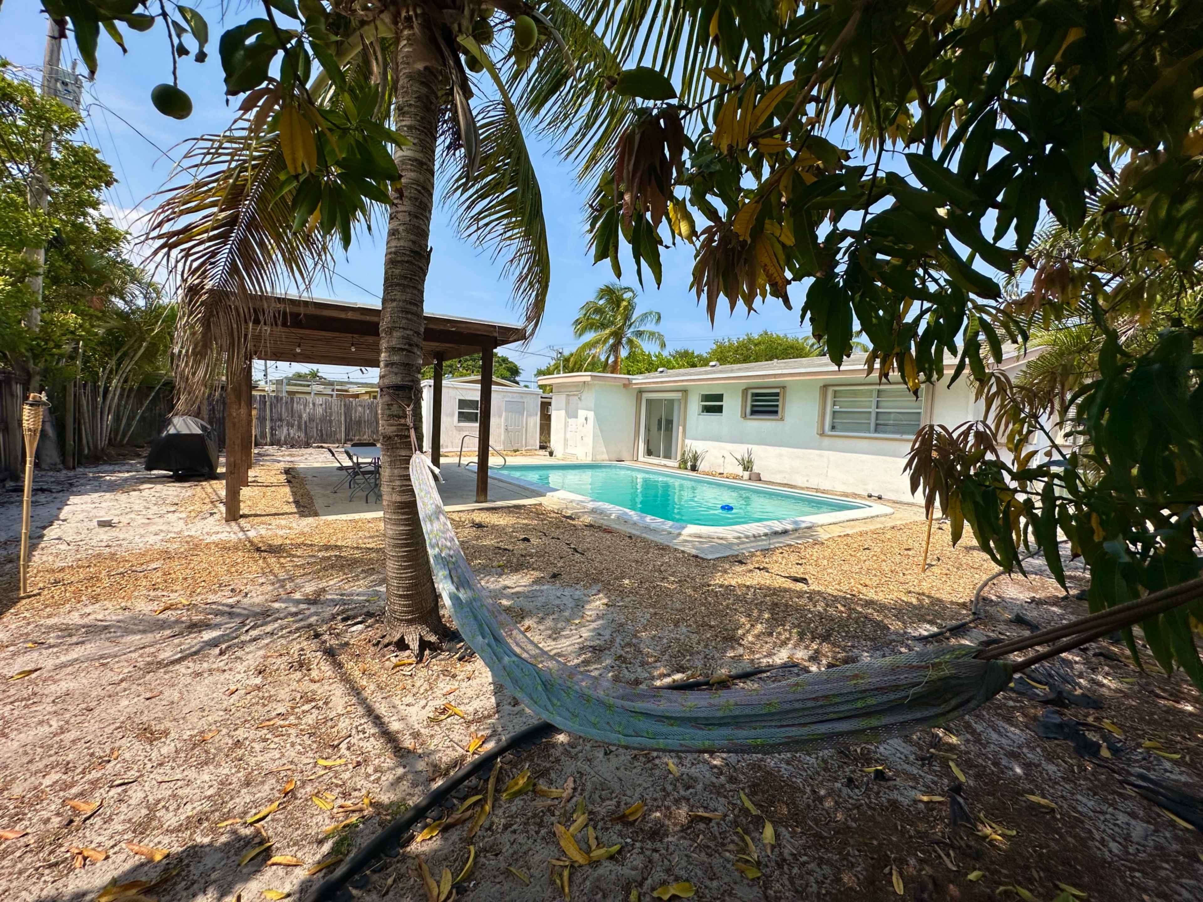 A backyard with a swimming pool, a hammock hanging between palm trees, and a wooden pergola beside a white house.