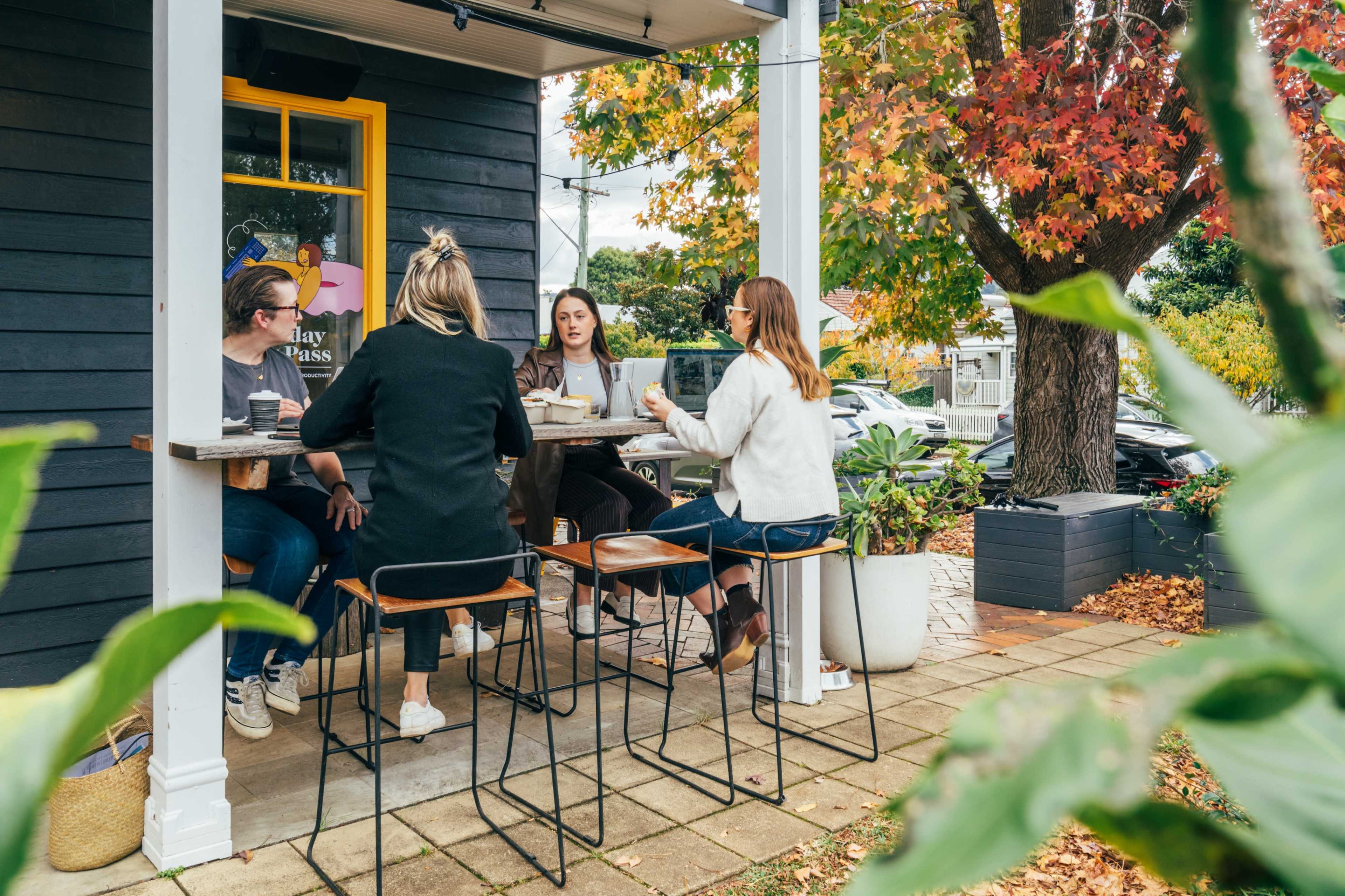 A group of five people sits at a table outside a coffee shop surrounded by autumn foliage.