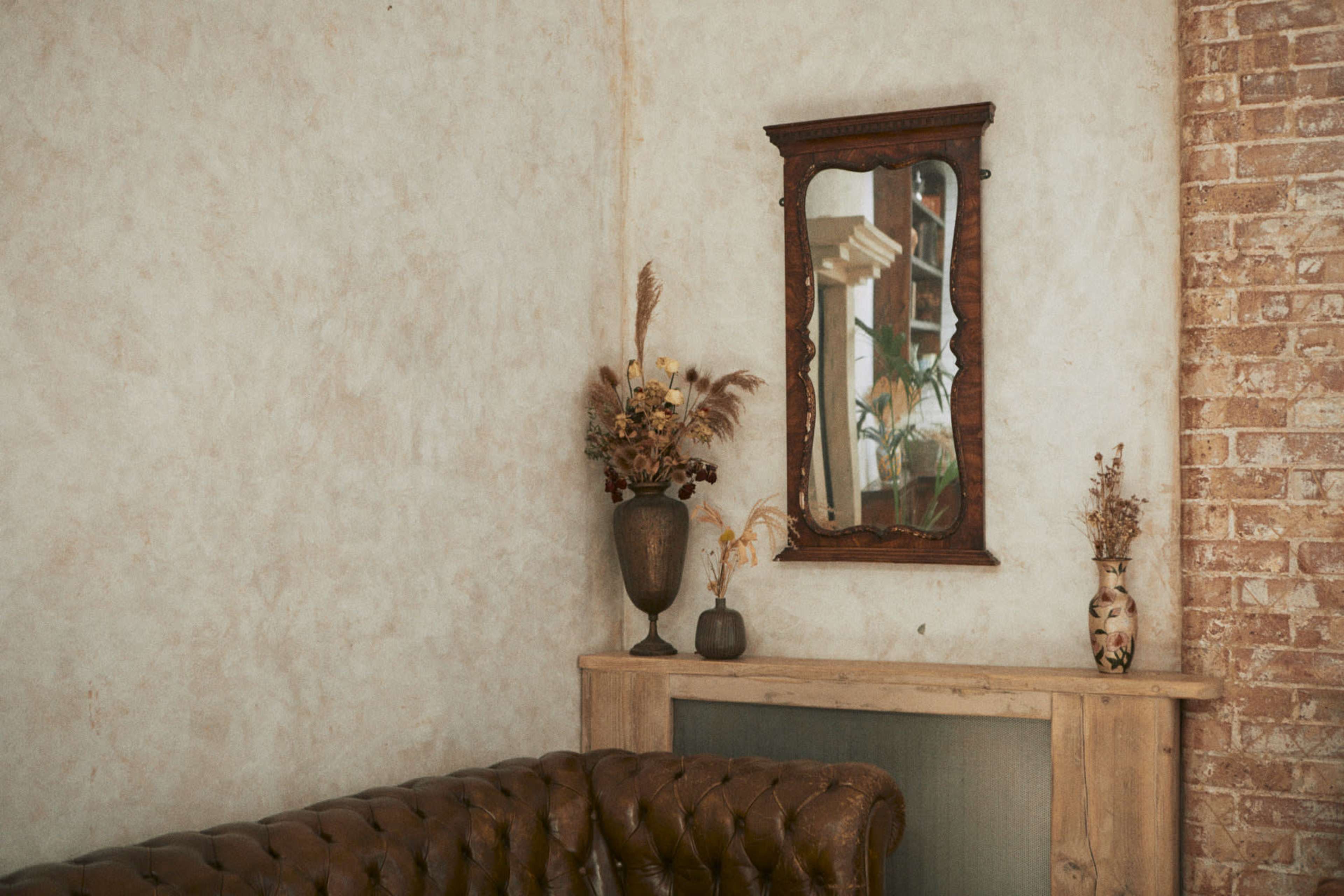 A corner of a room featuring a vintage leather sofa, a wooden table, and a wall-mounted mirror above decorative vases with dried flowers.