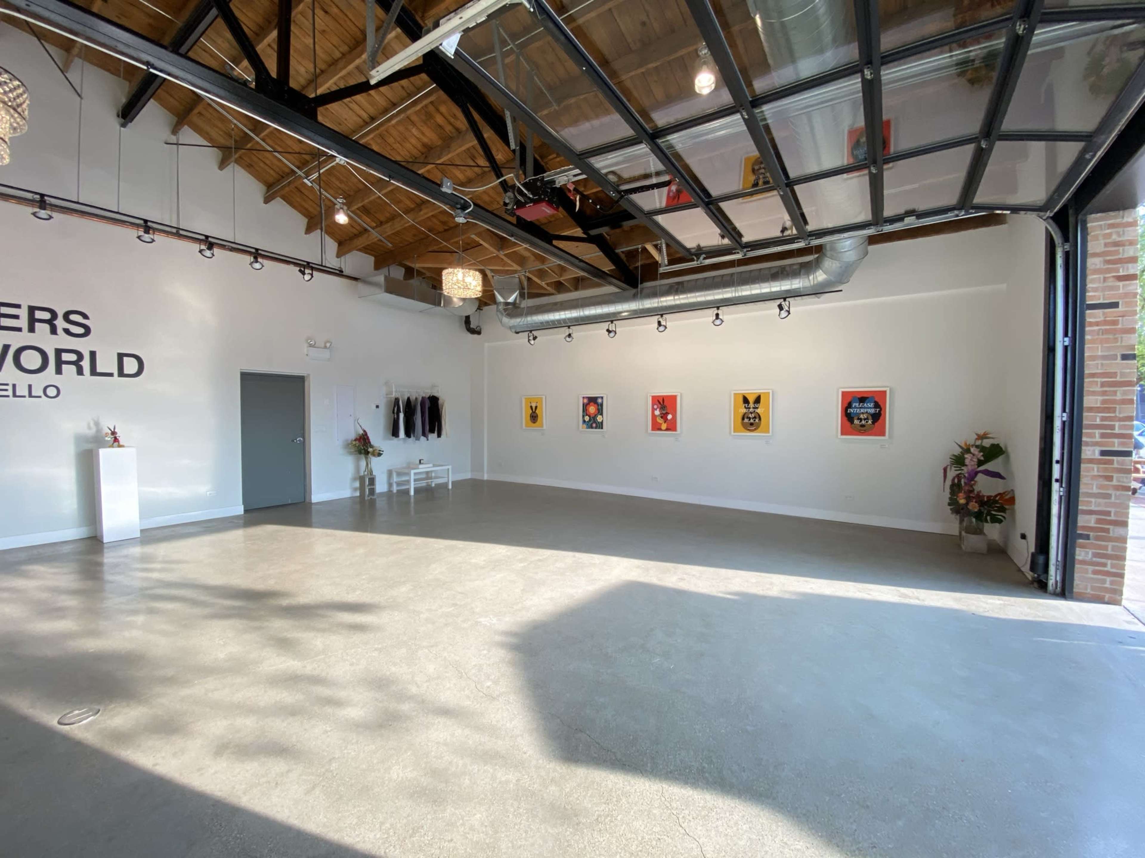 A spacious art gallery with polished concrete floors, white walls featuring framed artwork, and large glass doors leading outside.