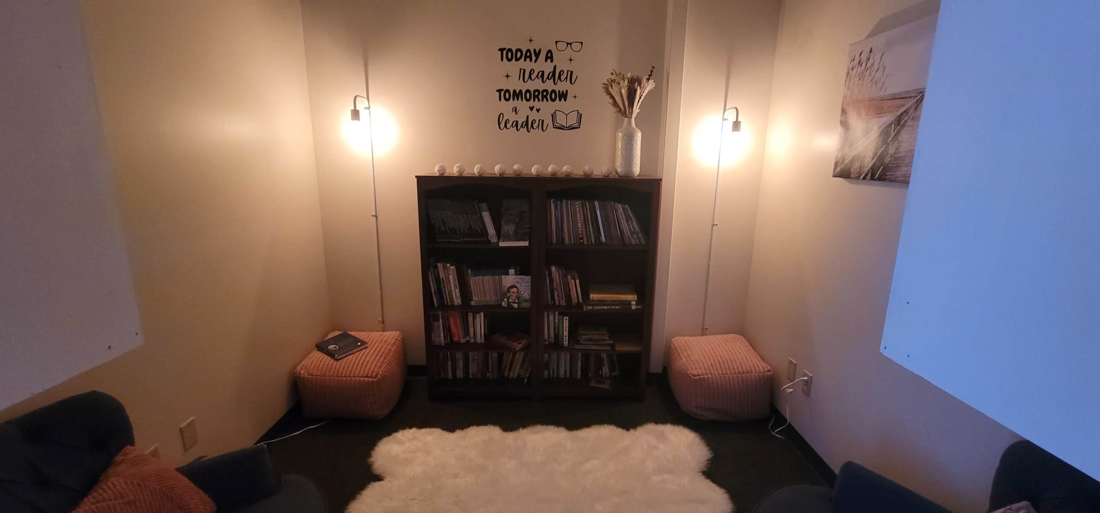 The image shows a cozy reading nook with a bookshelf, two poufs, a soft rug, and ambient lighting.