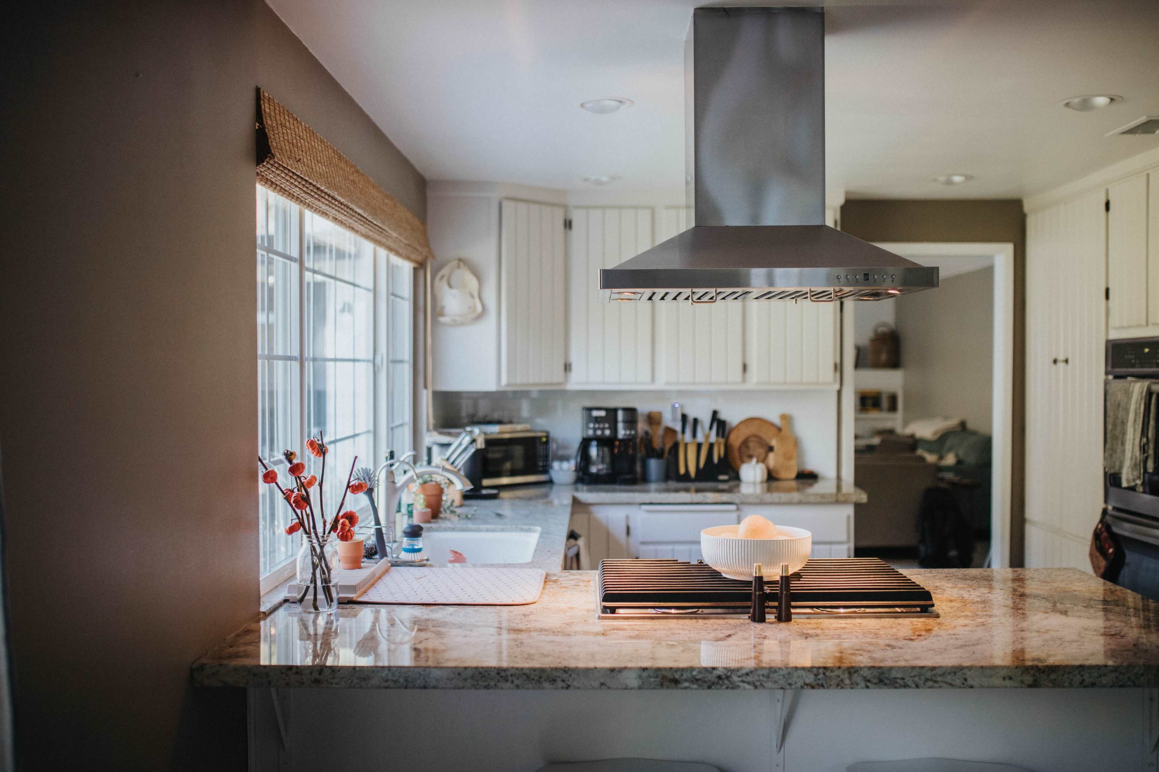 A bright kitchen features white cabinets, a central island with a stovetop, and large windows allowing natural light to enter.