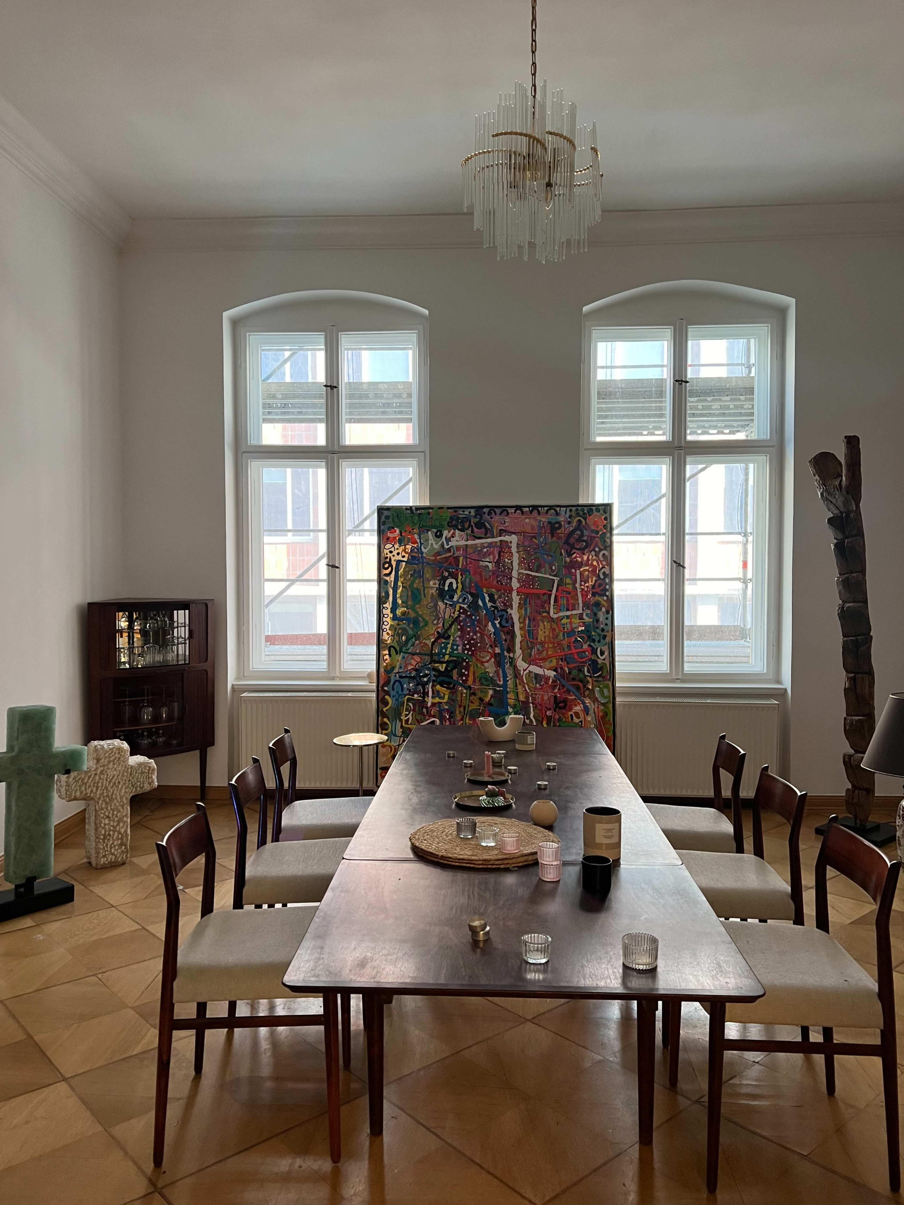 A long wooden dining table is set in a brightly lit room with large windows, featuring an abstract painting on an easel and various decorative items scattered across the table.